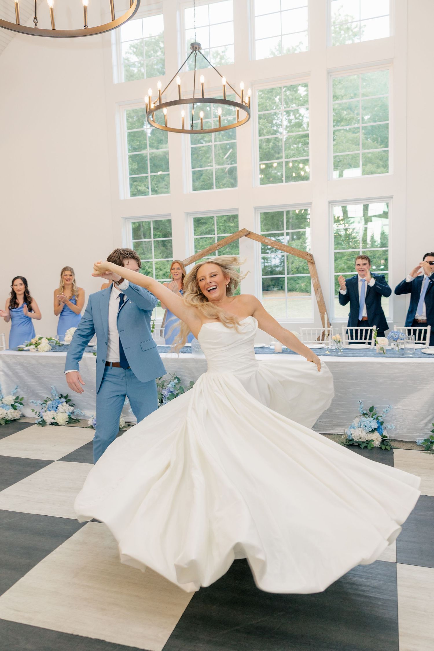 Bride twirls in white ballgown as groom spins her during first dance in bright elegant reception hall.