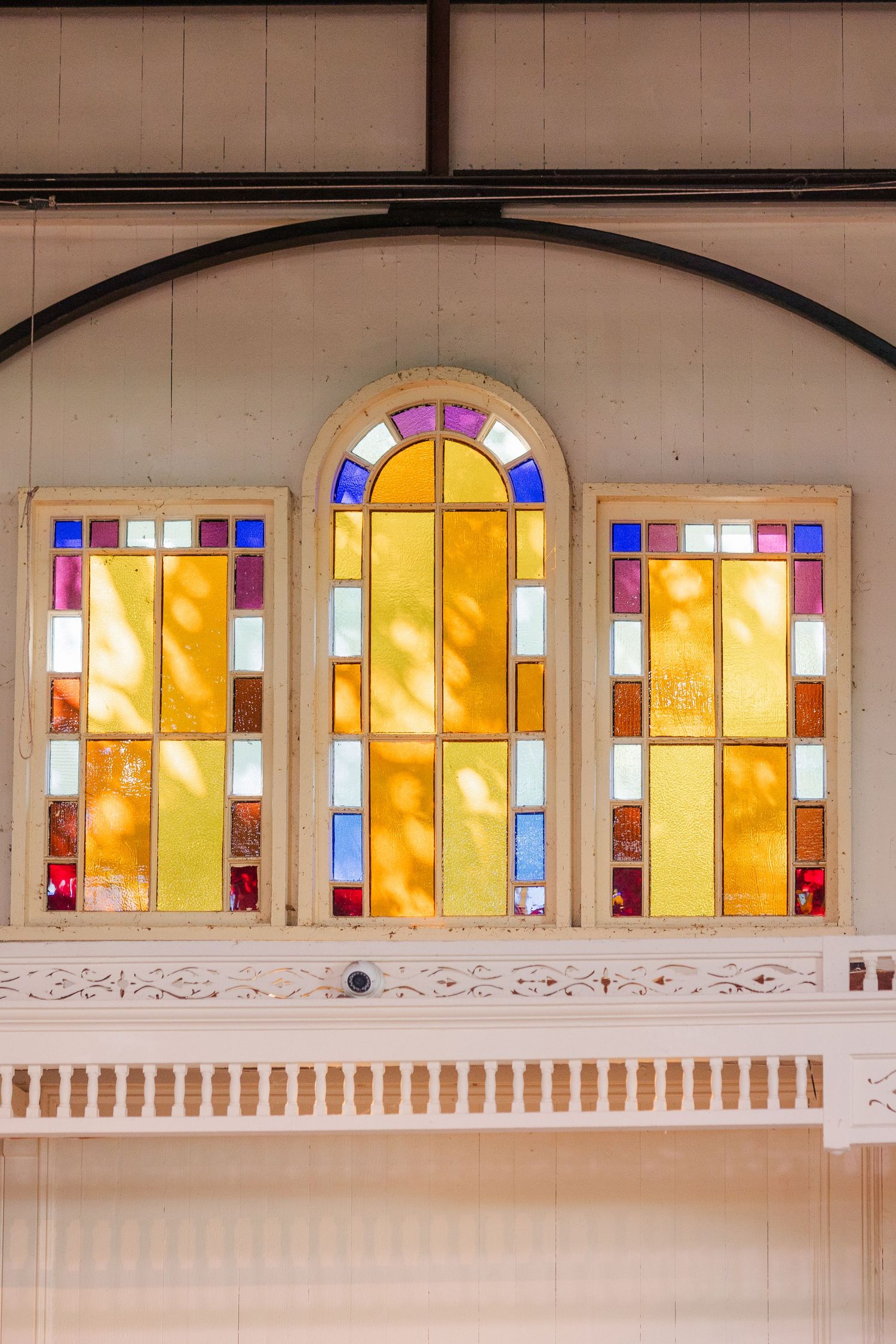 Three colorful stained glass windows with arched center panel in a white wooden church interior.
