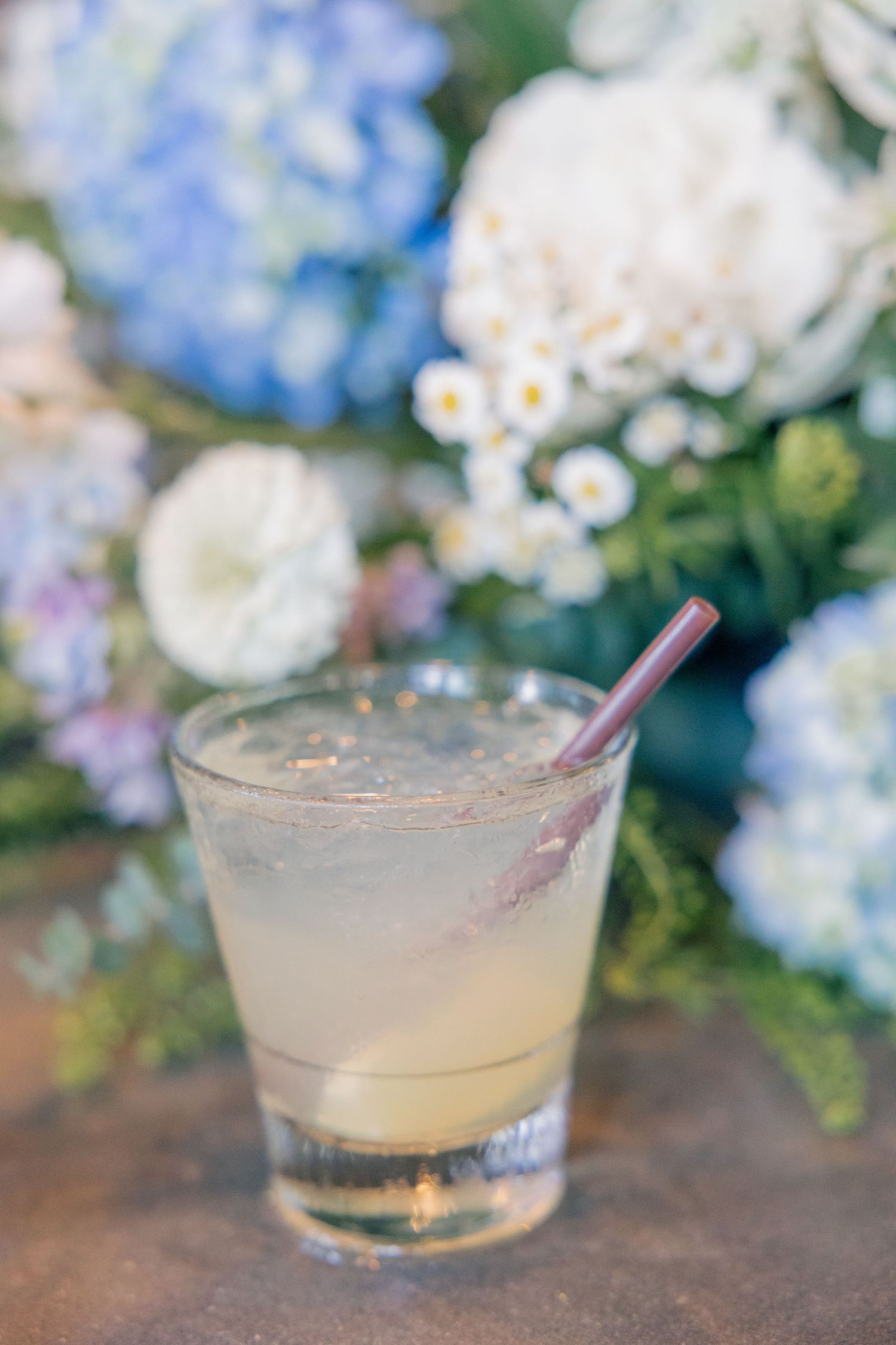 A refreshing lemonade cocktail with a pink straw in a glass surrounded by blue and white hydrangea flowers.