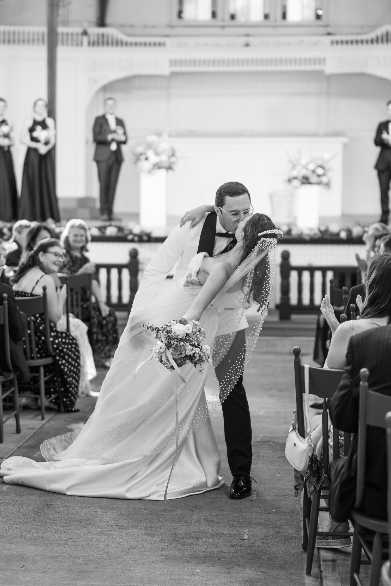 Black and white photo of groom dipping bride for kiss while walking down church aisle after ceremony.