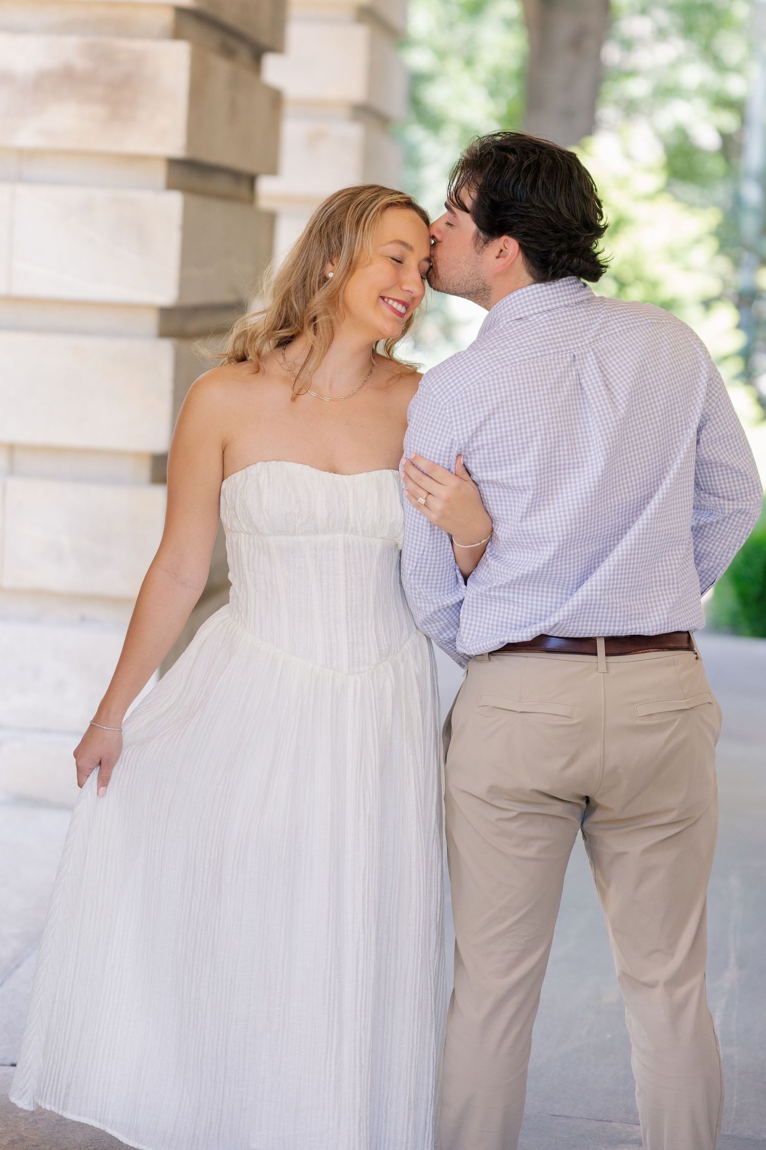 A groom kisses his smiling bride on the cheek as she wears a strapless white dress outdoors.