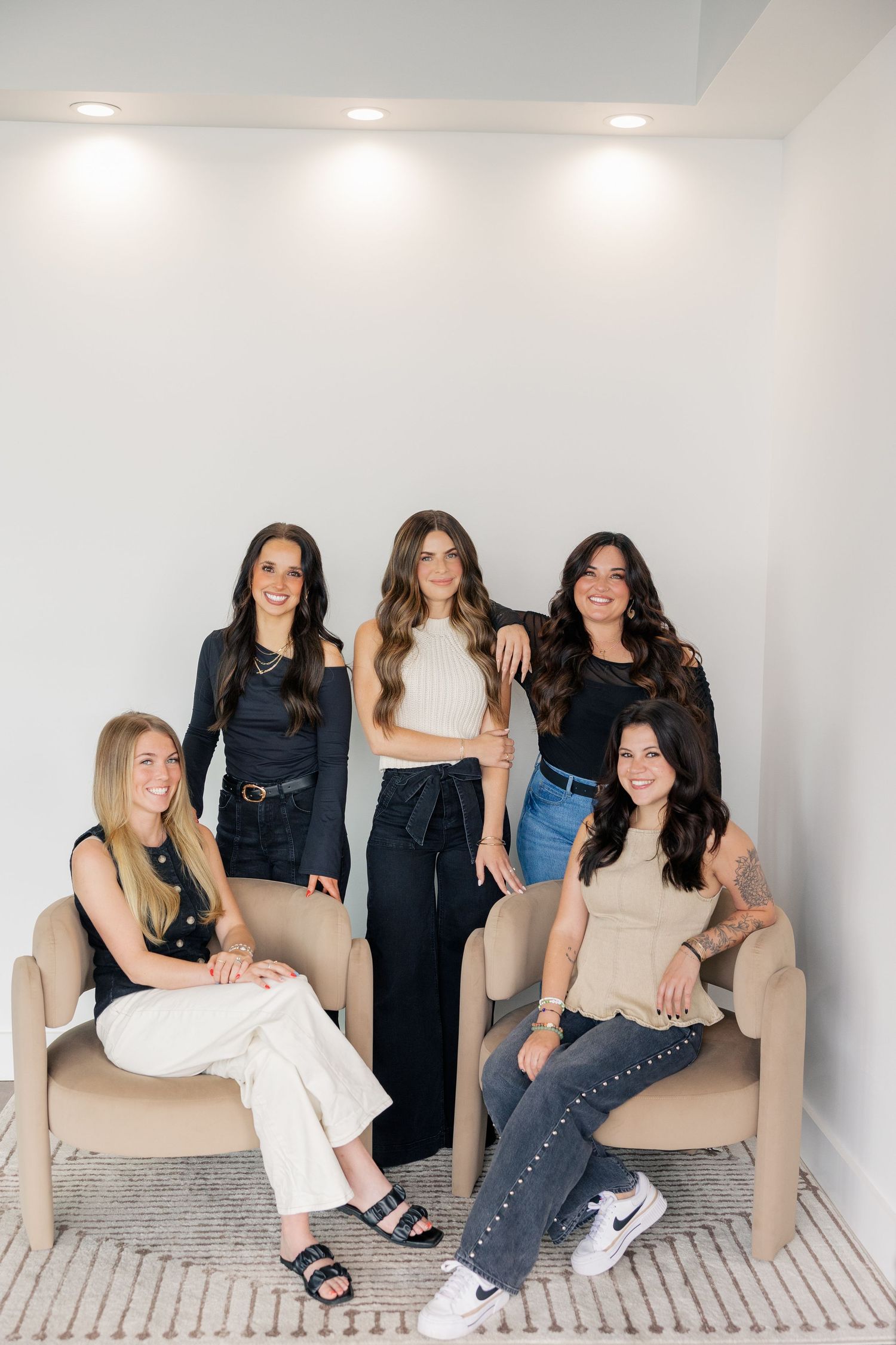 Five women pose together in a modern studio, three standing and two seated in beige chairs, wearing stylish casual outfits.