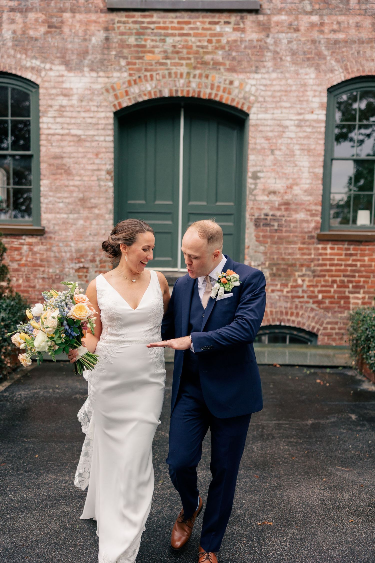 Bride and groom walk together outside a historic brick building, bride holding a beautiful peach and white bouquet.