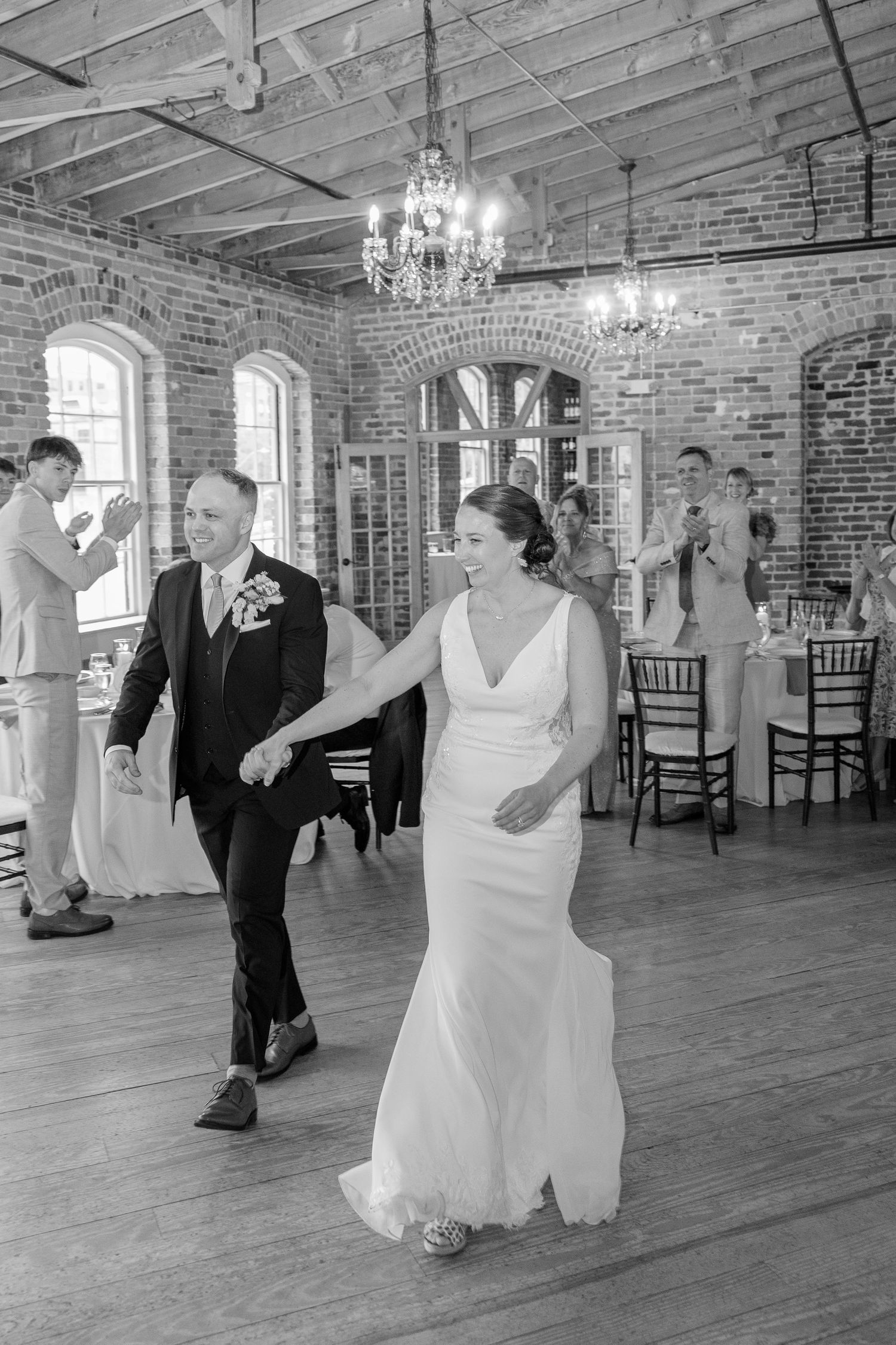 Bride and groom make grand entrance into rustic brick reception hall with chandeliers in black and white.