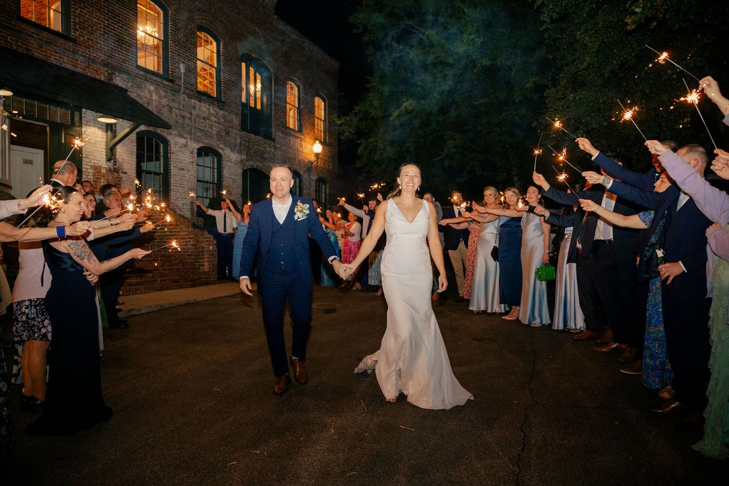 Bride and groom exit wedding reception through sparkler tunnel surrounded by cheering guests at night.