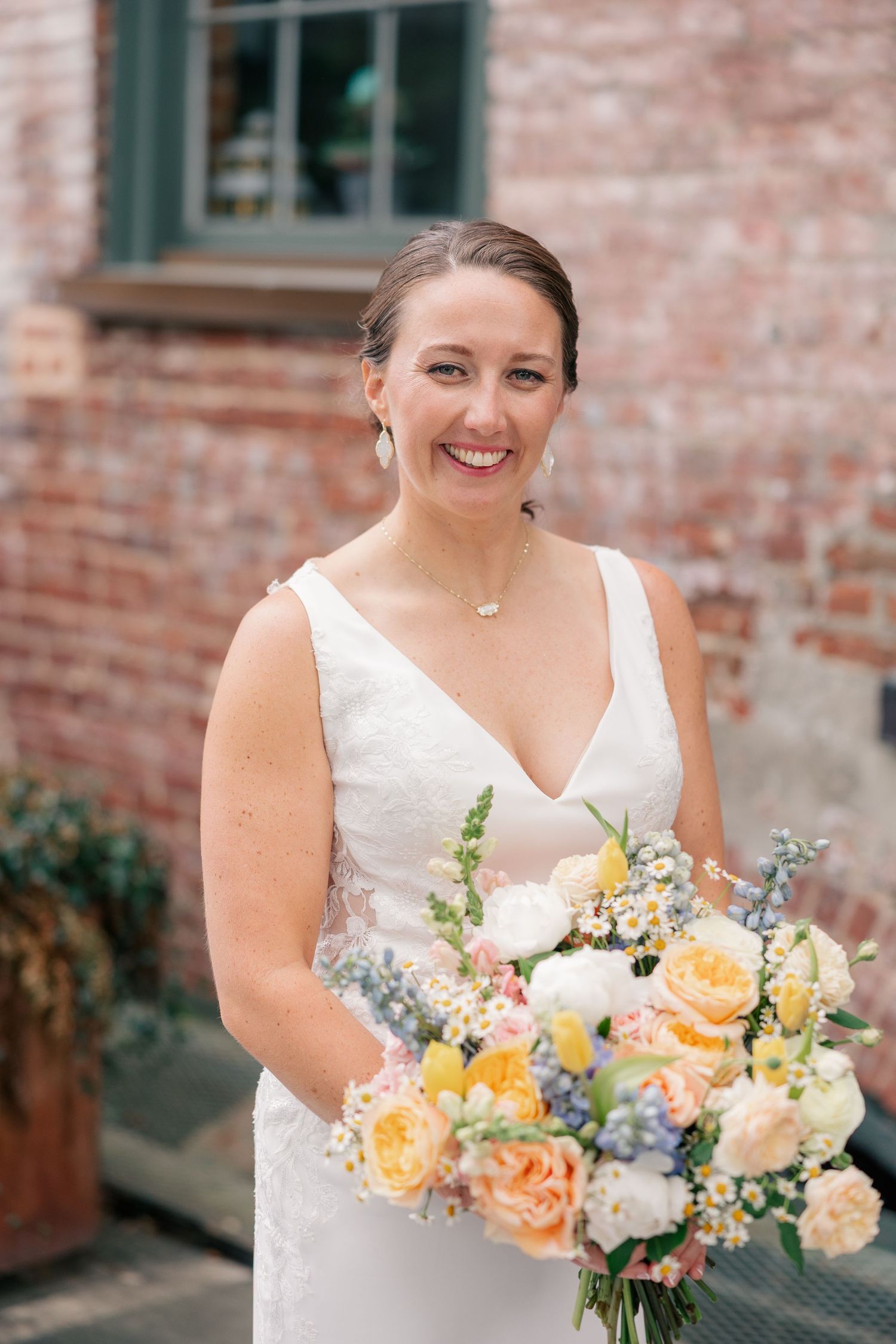 Smiling bride in white V-neck gown holds colorful spring bouquet with roses and wildflowers.