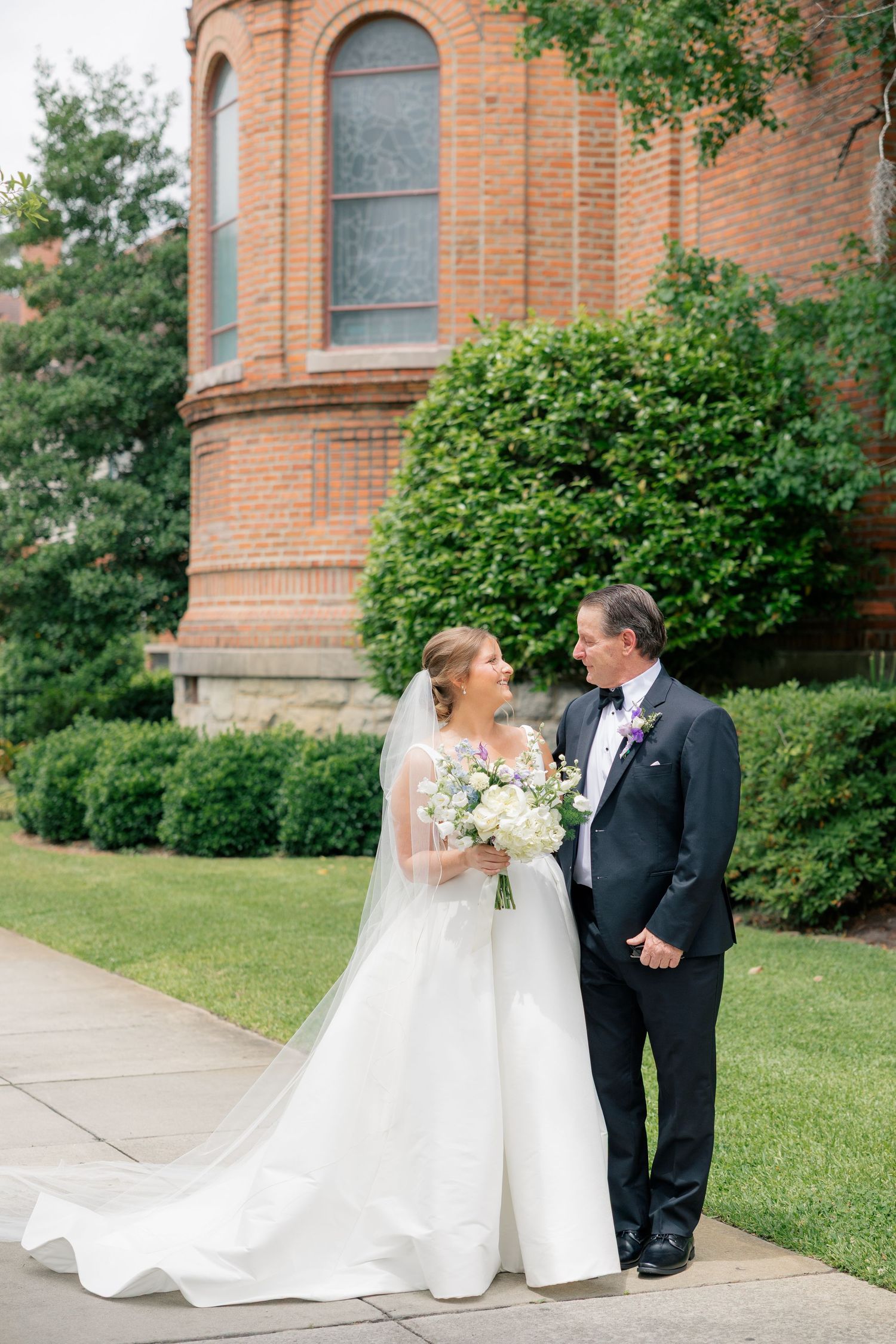 Bride and groom standing together outside brick church, bride holding white floral bouquet with long cathedral veil.