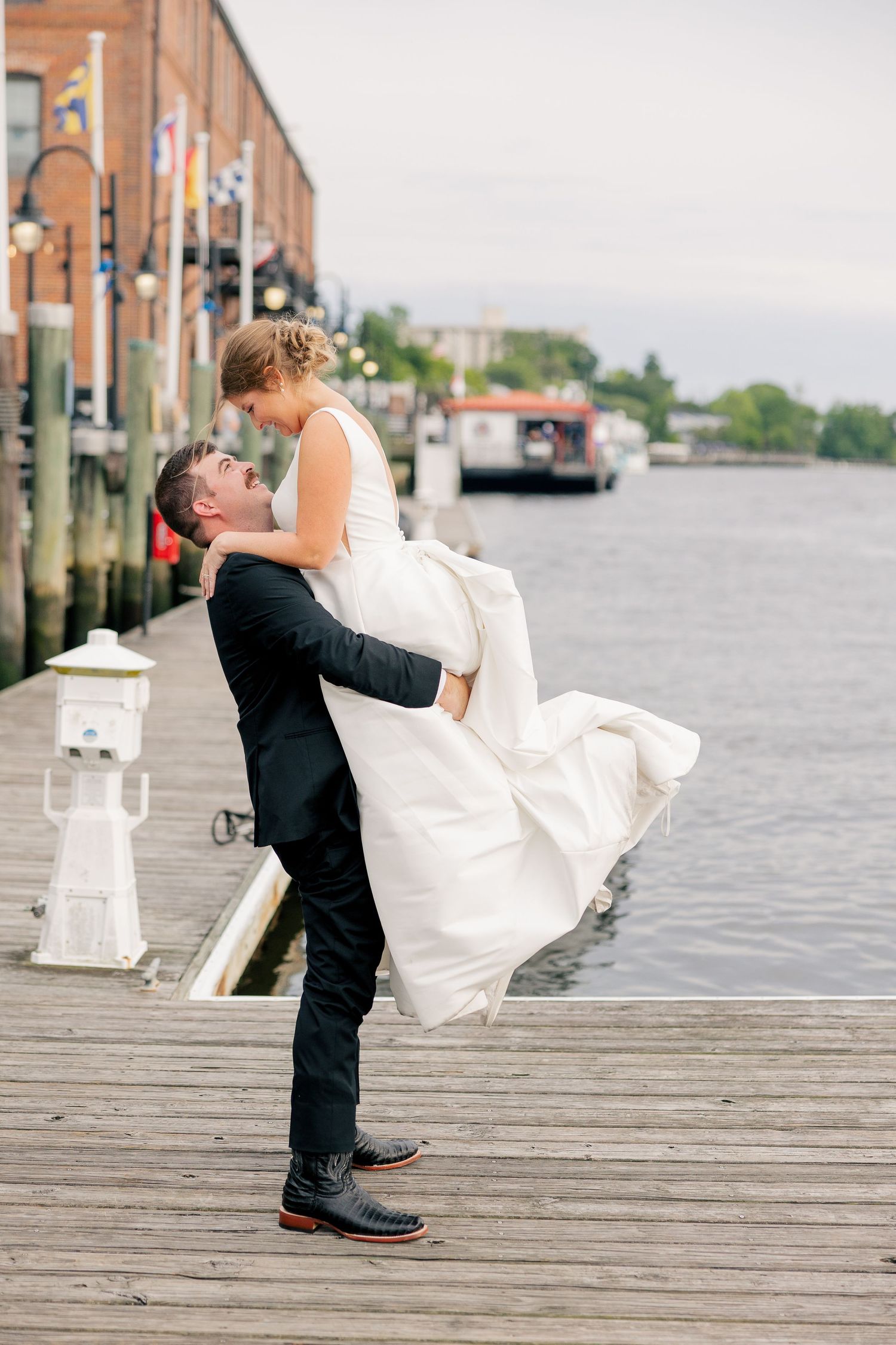 Groom lifting bride in white dress on waterfront dock at wedding.