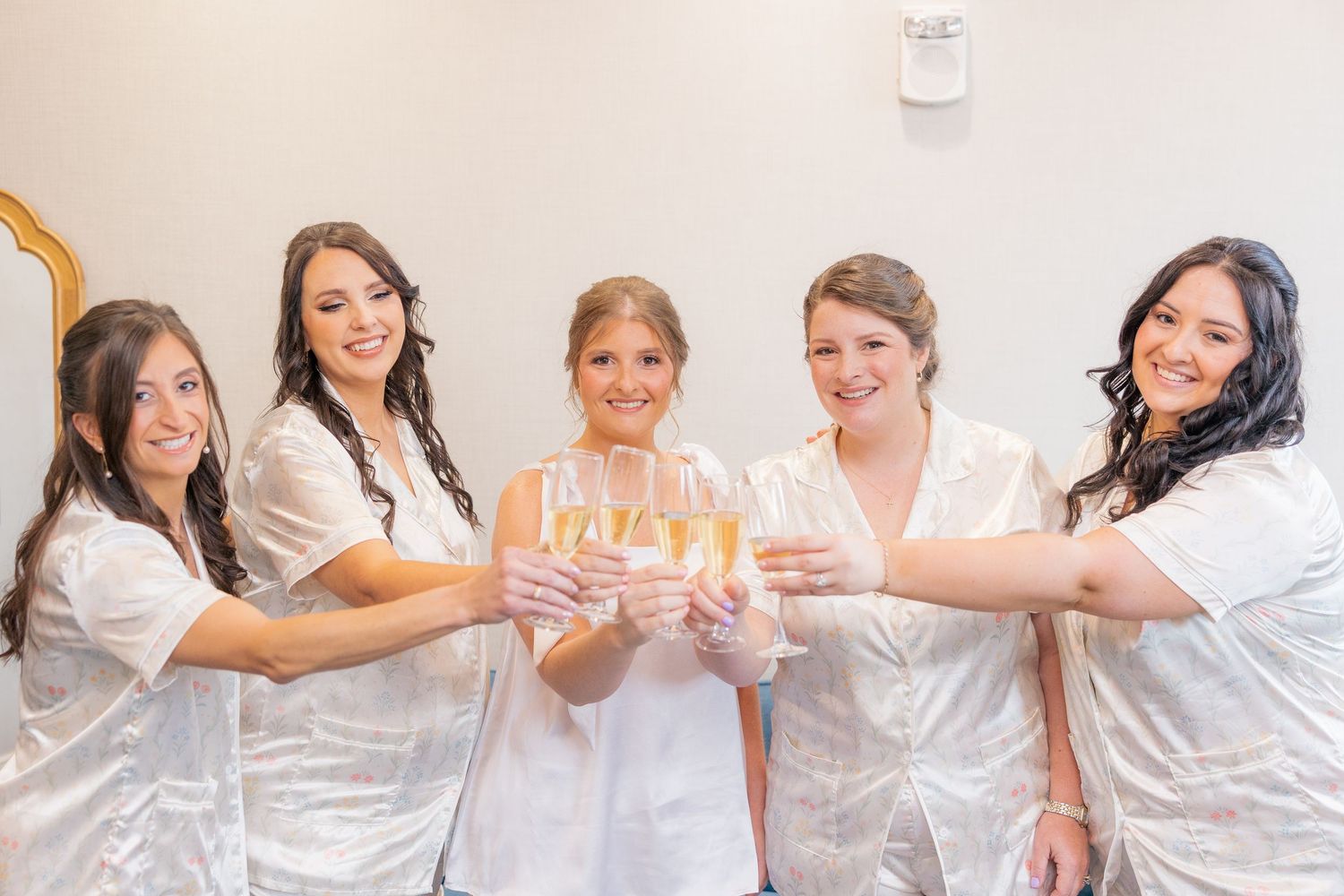 Five bridesmaids in white robes toasting with champagne glasses, smiling together before a wedding celebration.