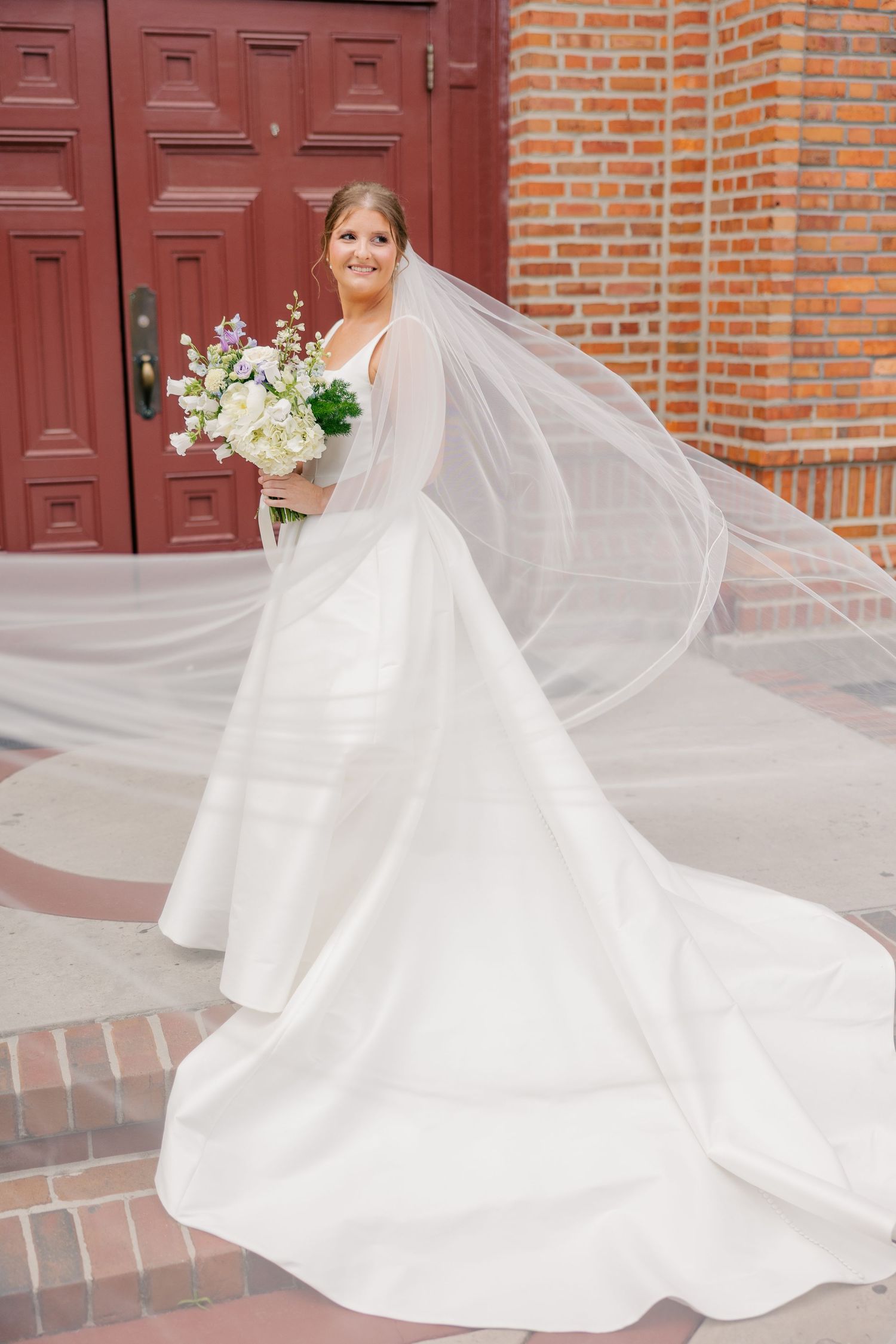 Bride in flowing white gown and veil holding bouquet outside red brick church.