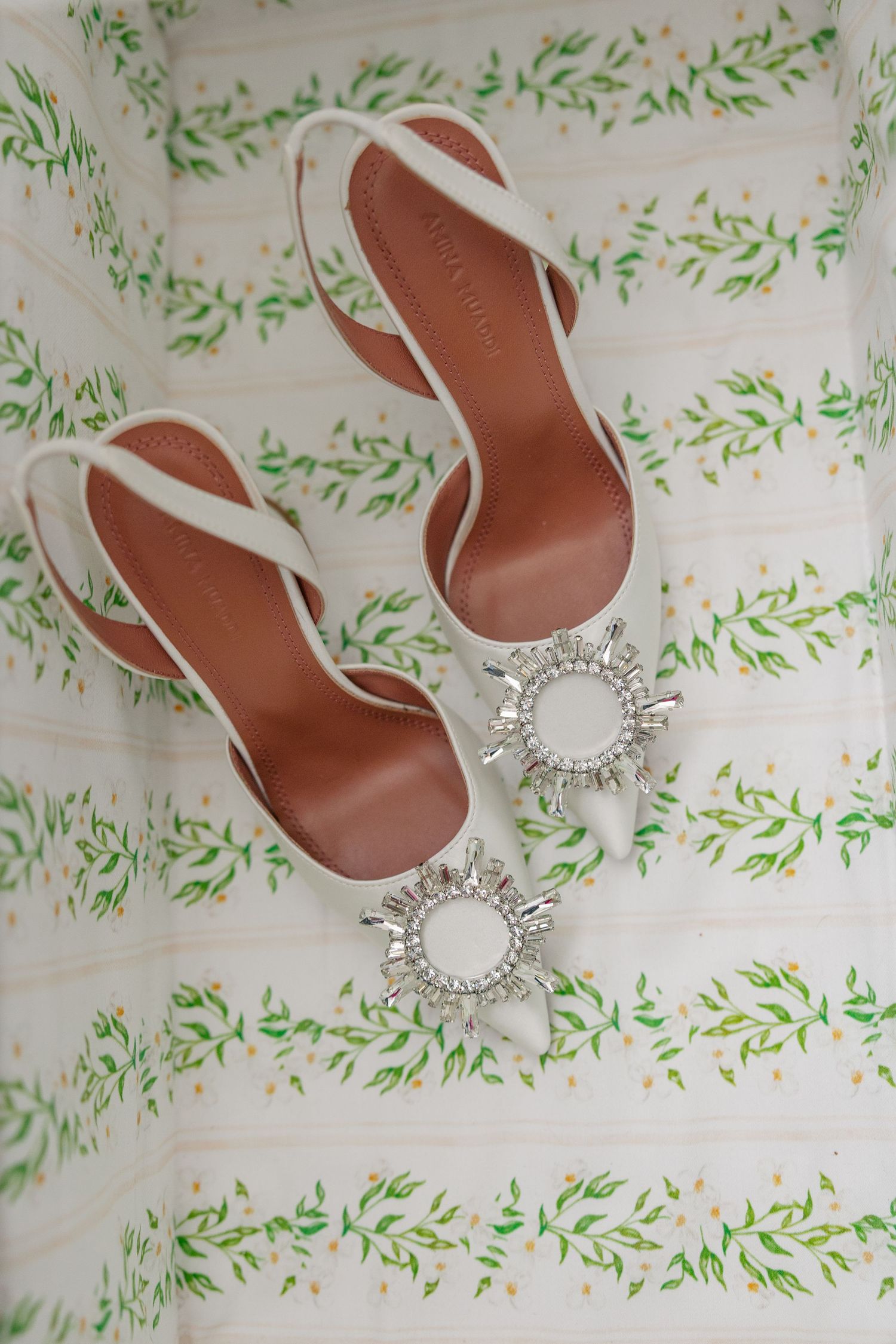 White bridal heeled sandals with crystal sunburst embellishments displayed on a floral green and white fabric.