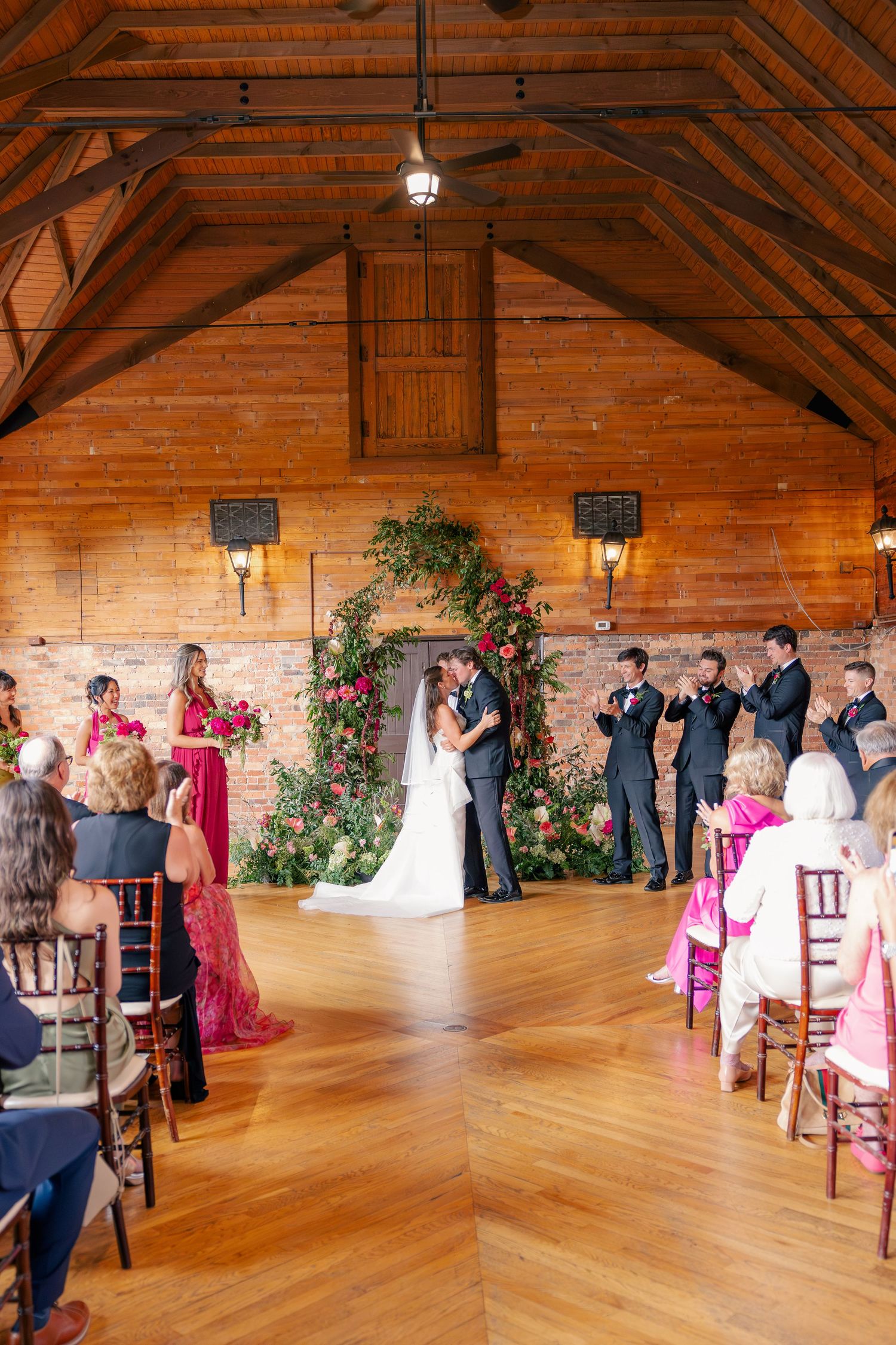 Couple shares first kiss at altar beneath lush floral arch in elegant wooden barn ceremony venue.