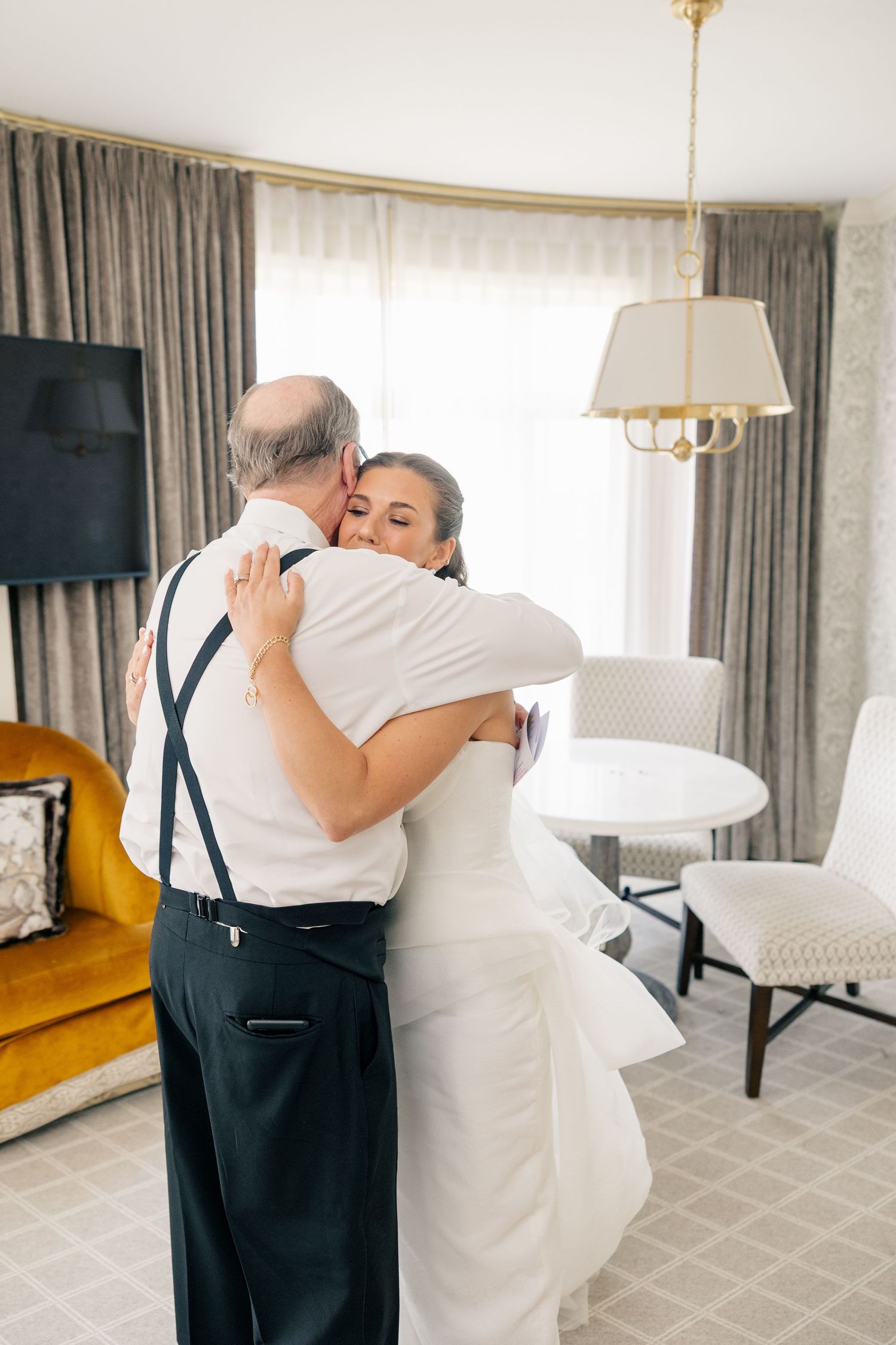 Bride in white gown shares a warm embrace with her father in a hotel room.