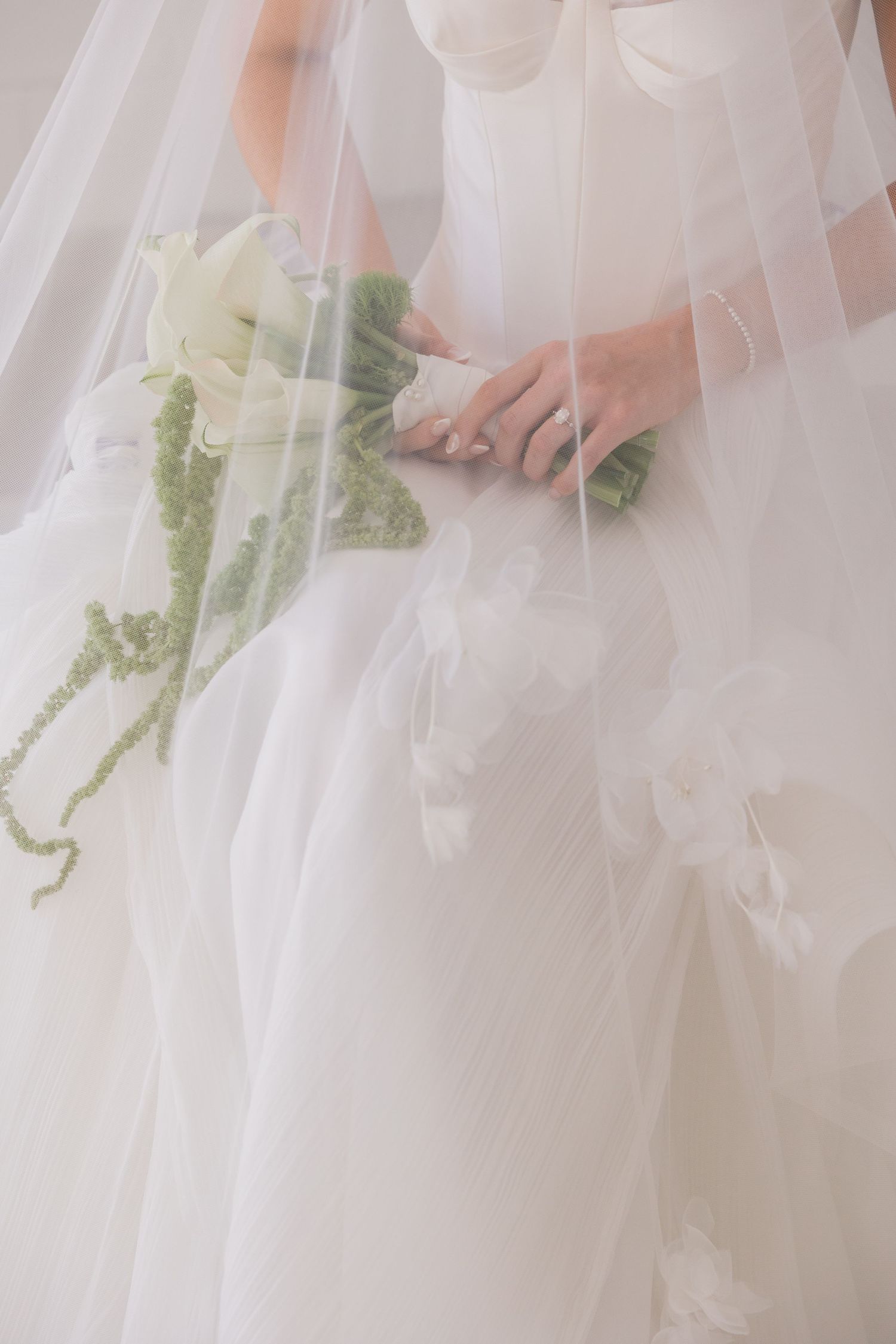 Delicate white bridal veil drapes over hands holding an elegant bouquet with white flowers and green foliage.