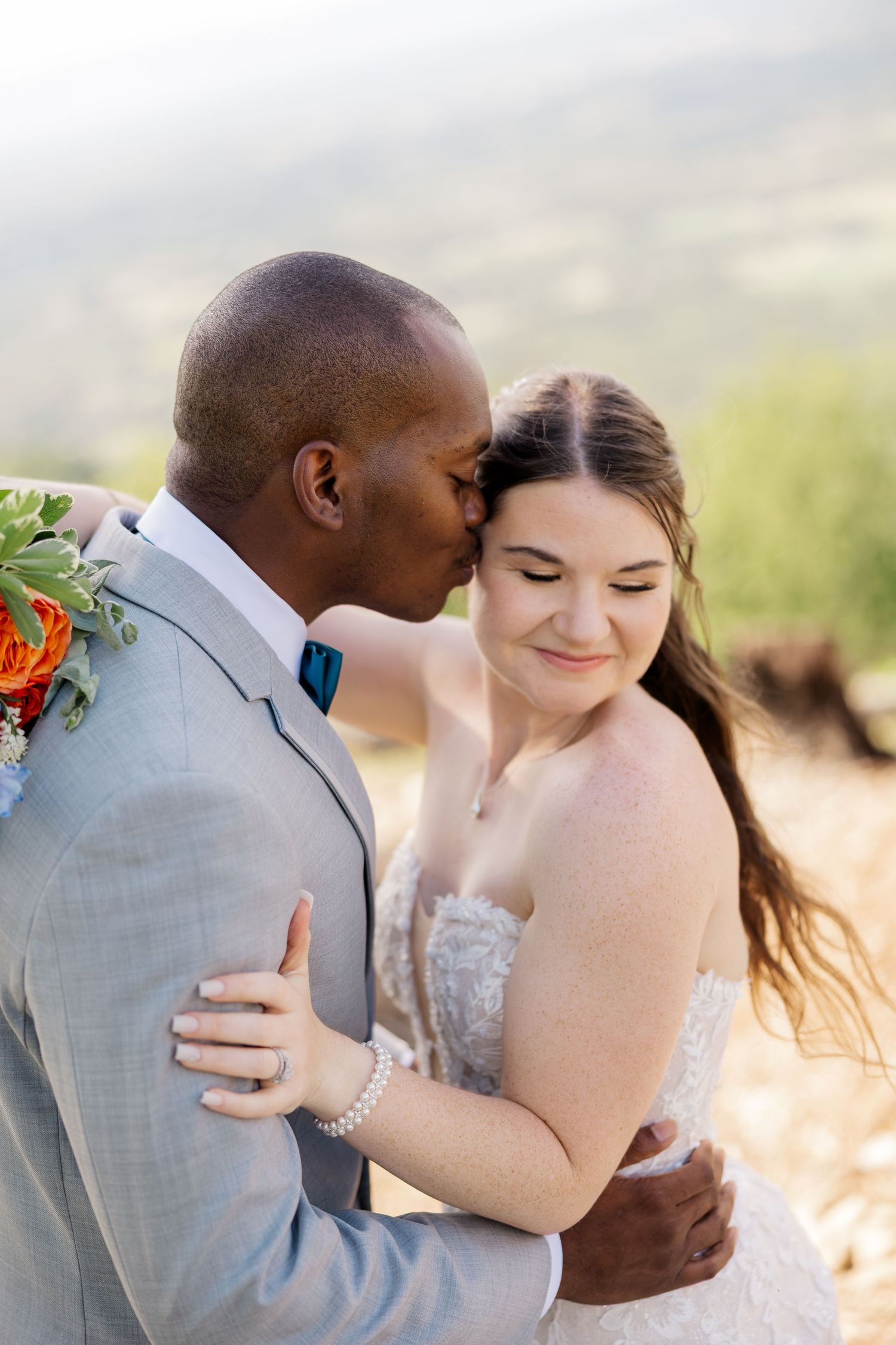 A groom in a gray suit kisses his bride on the forehead during an outdoor wedding ceremony.