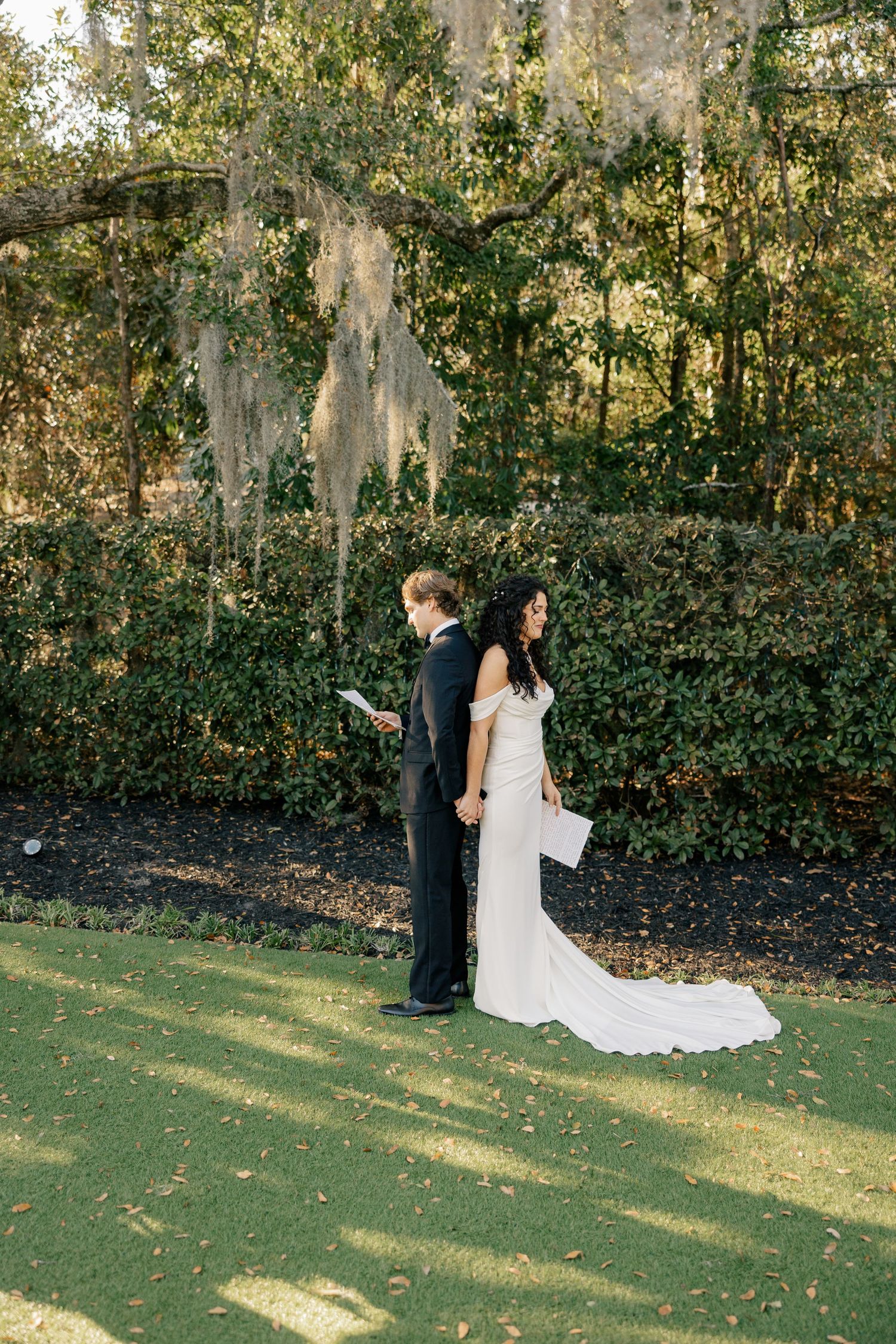 A couple in wedding attire poses back-to-back while reading letters outdoors beneath Spanish moss.