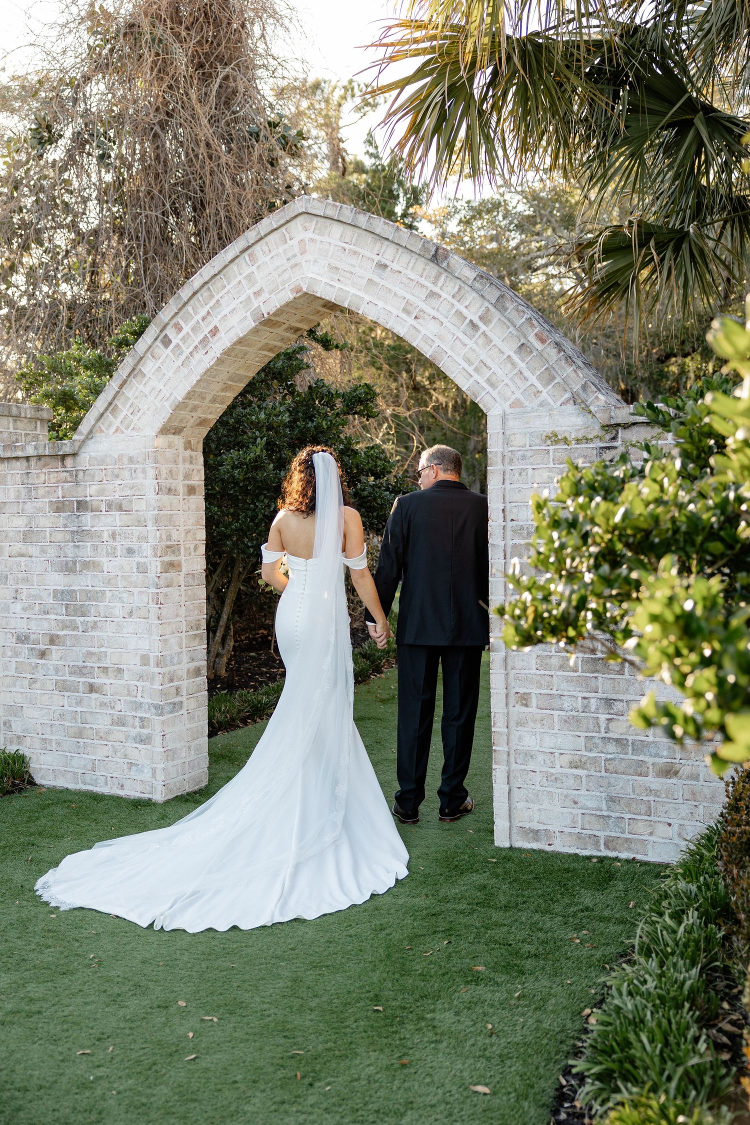 A wedding couple stands before an elegant stone archway surrounded by lush greenery and palm trees on a manicured lawn.