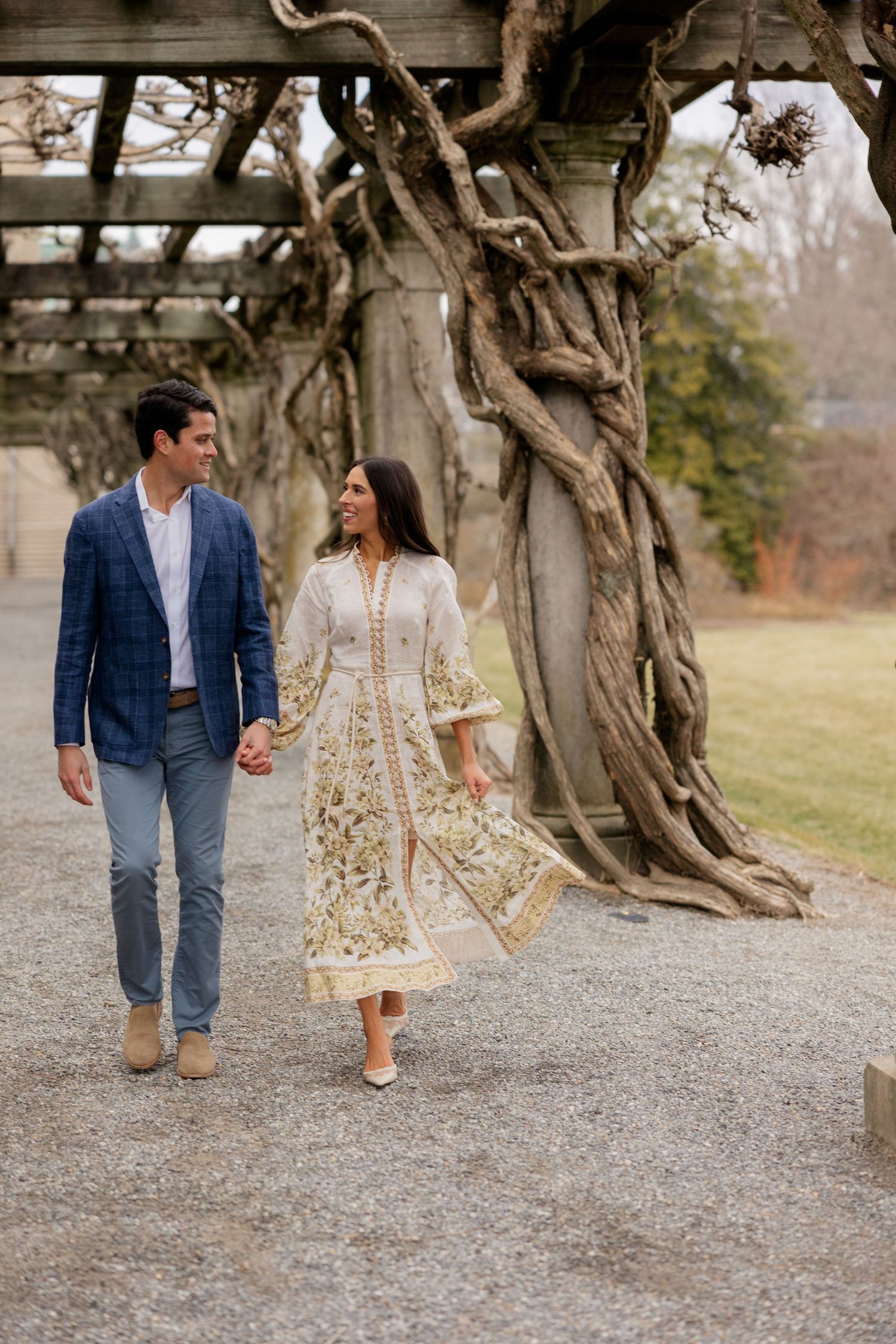 A couple walks hand in hand along a garden path beneath twisting vines on an elegant pergola arch structure.