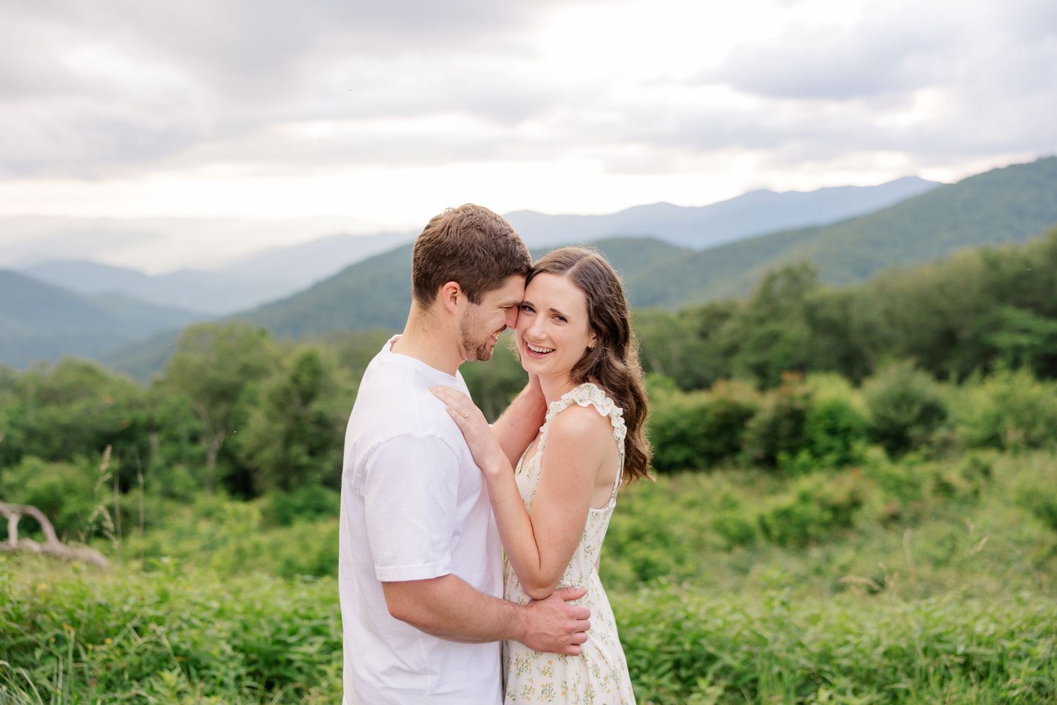 A couple shares tender moments in a scenic mountain meadow with lush greenery and rolling hills in the background.