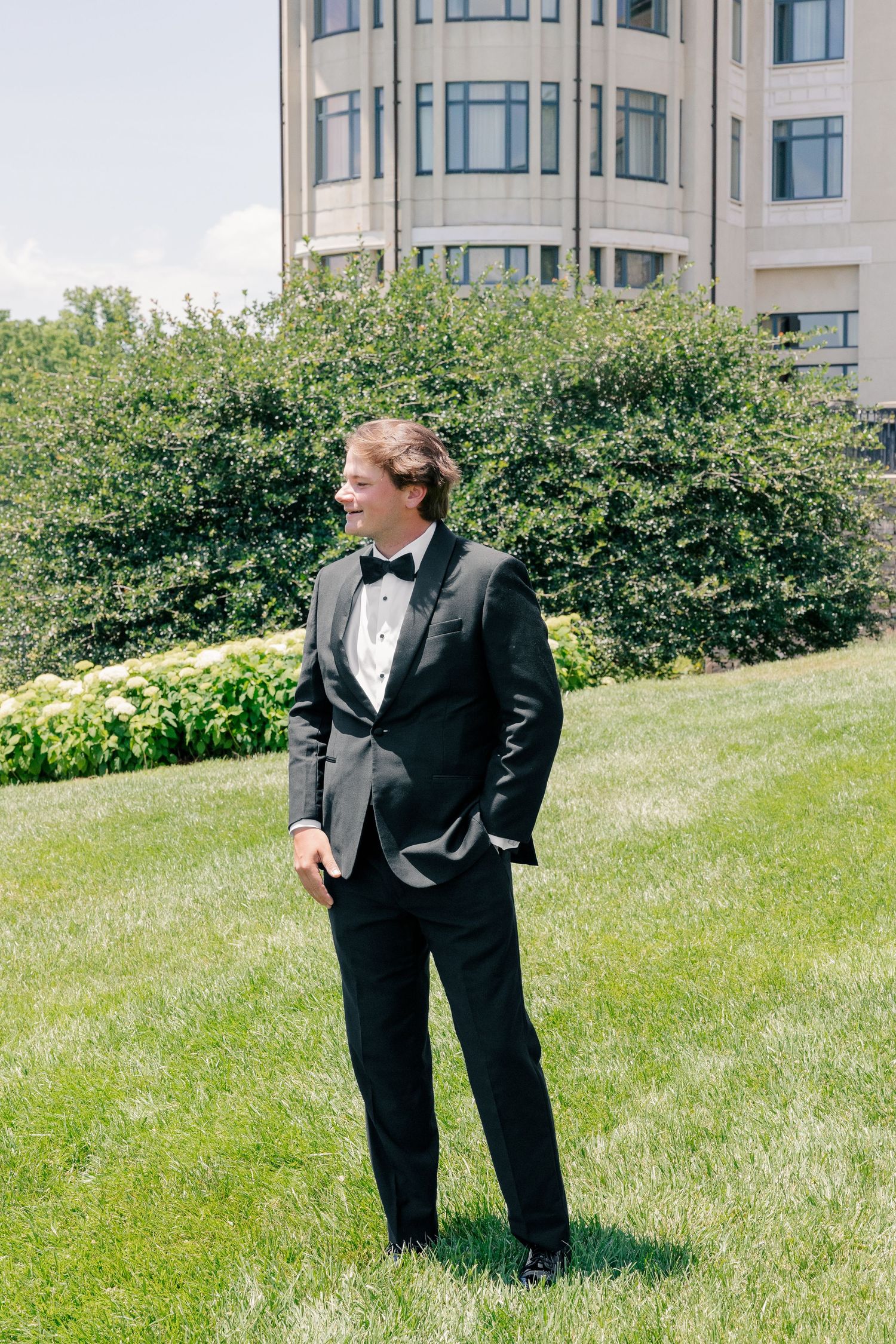 Groom in black tuxedo with bow tie stands on green lawn outside a large building on sunny day.