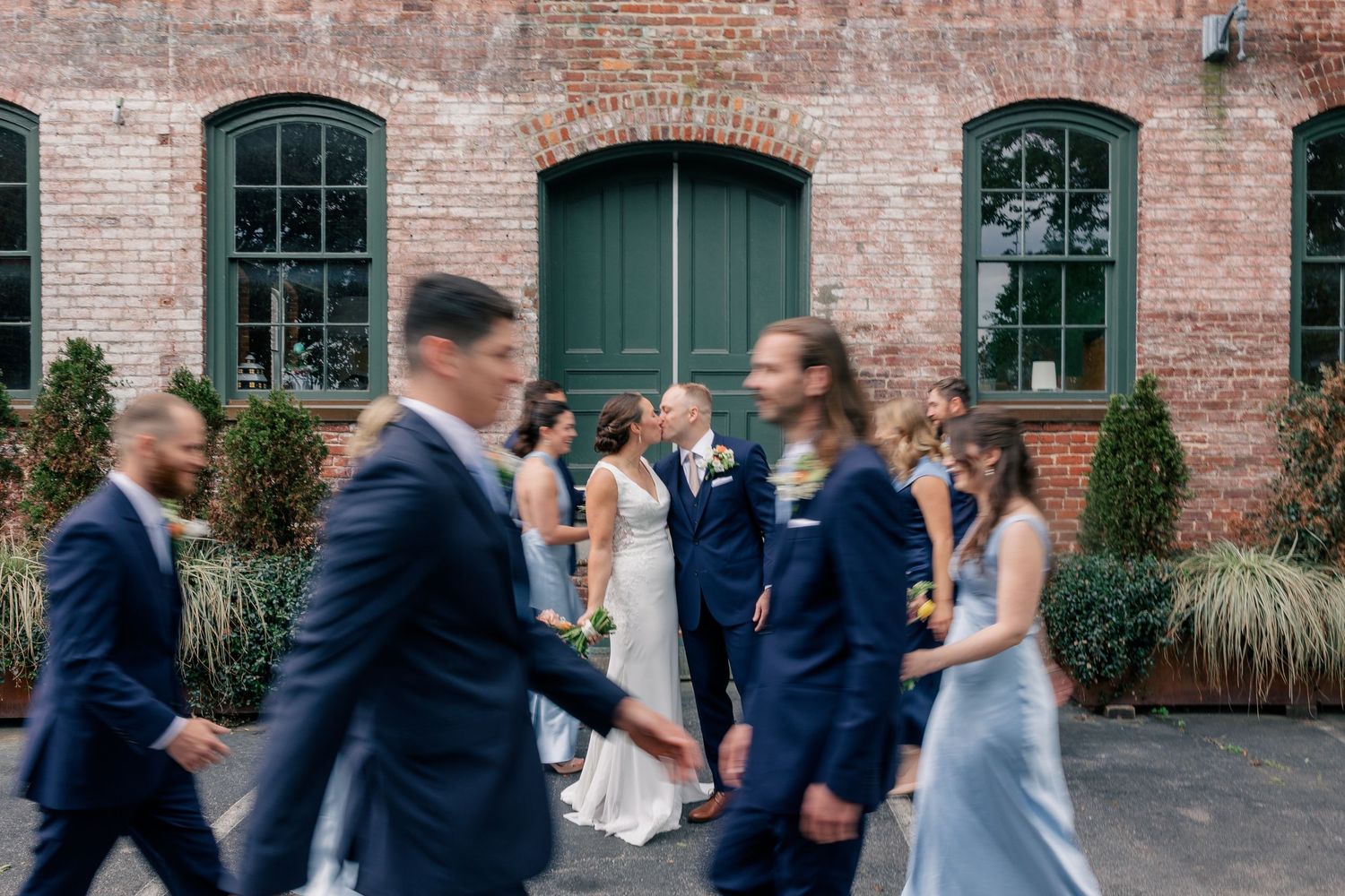 Wedding party walks past kissing bride and groom in a motion-blur photo at brick building venue.
