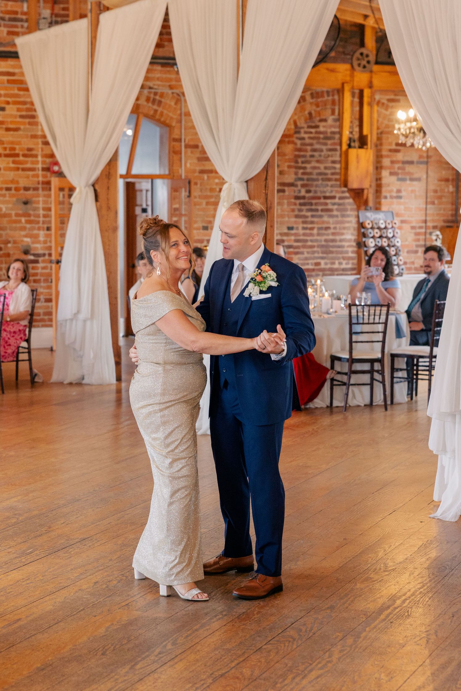 Groom in navy suit shares mother-son dance in elegant rustic brick reception hall with draping.
