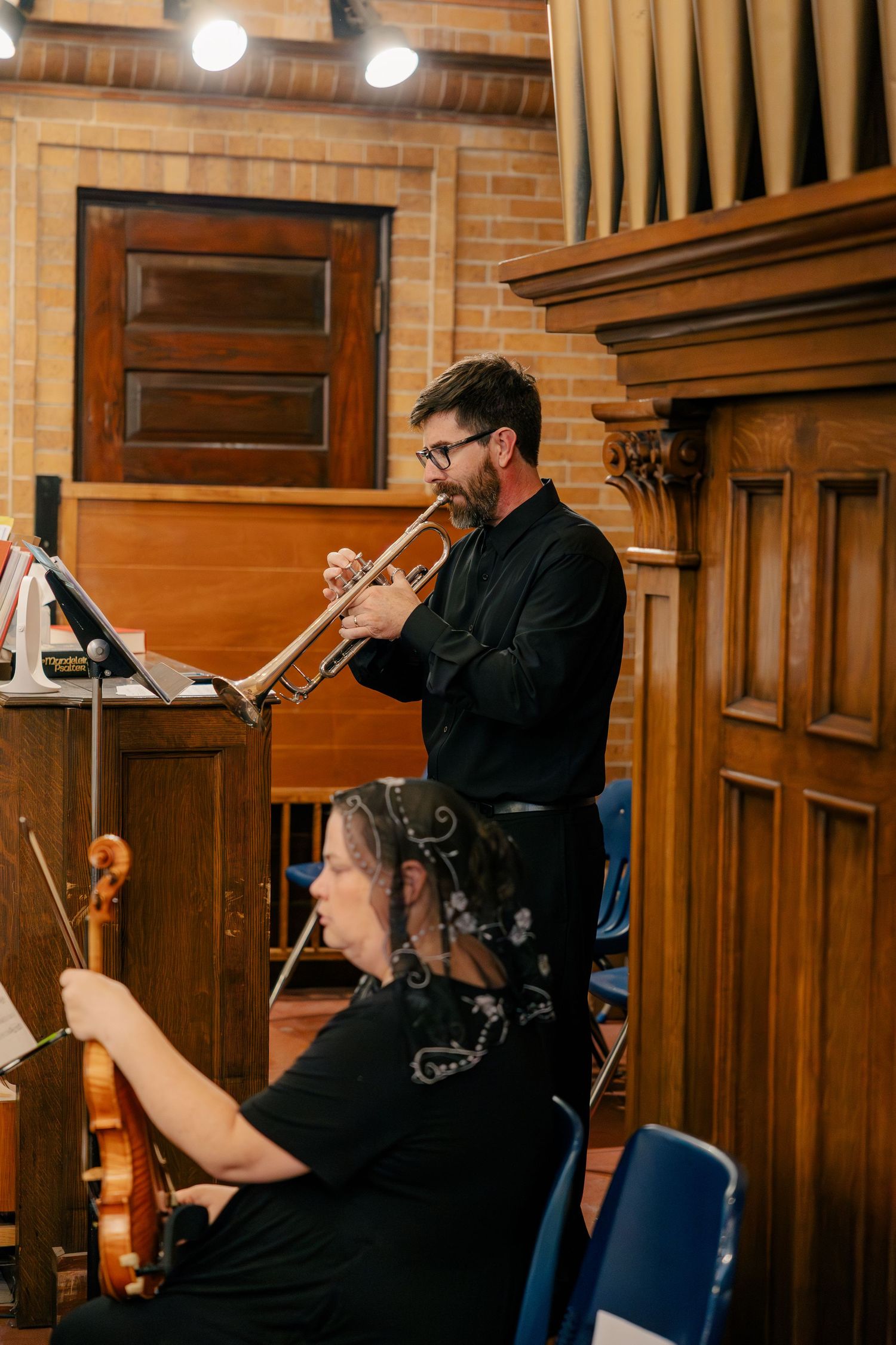 Musician playing trumpet beside a seated violinist during a wedding ceremony in a wooden church interior.