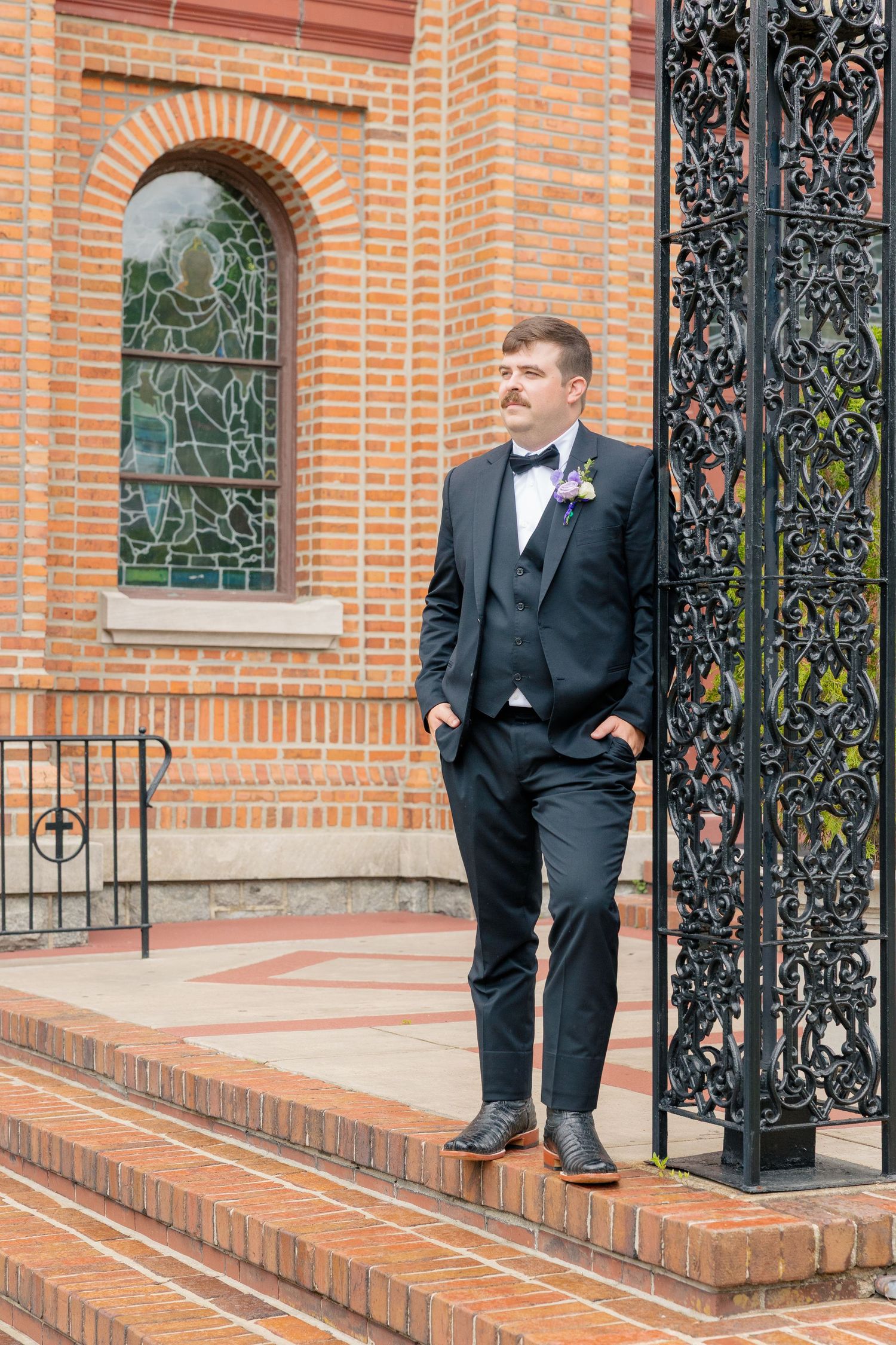 Groom in black tuxedo leaning against ornate iron gate outside brick church with stained glass window.