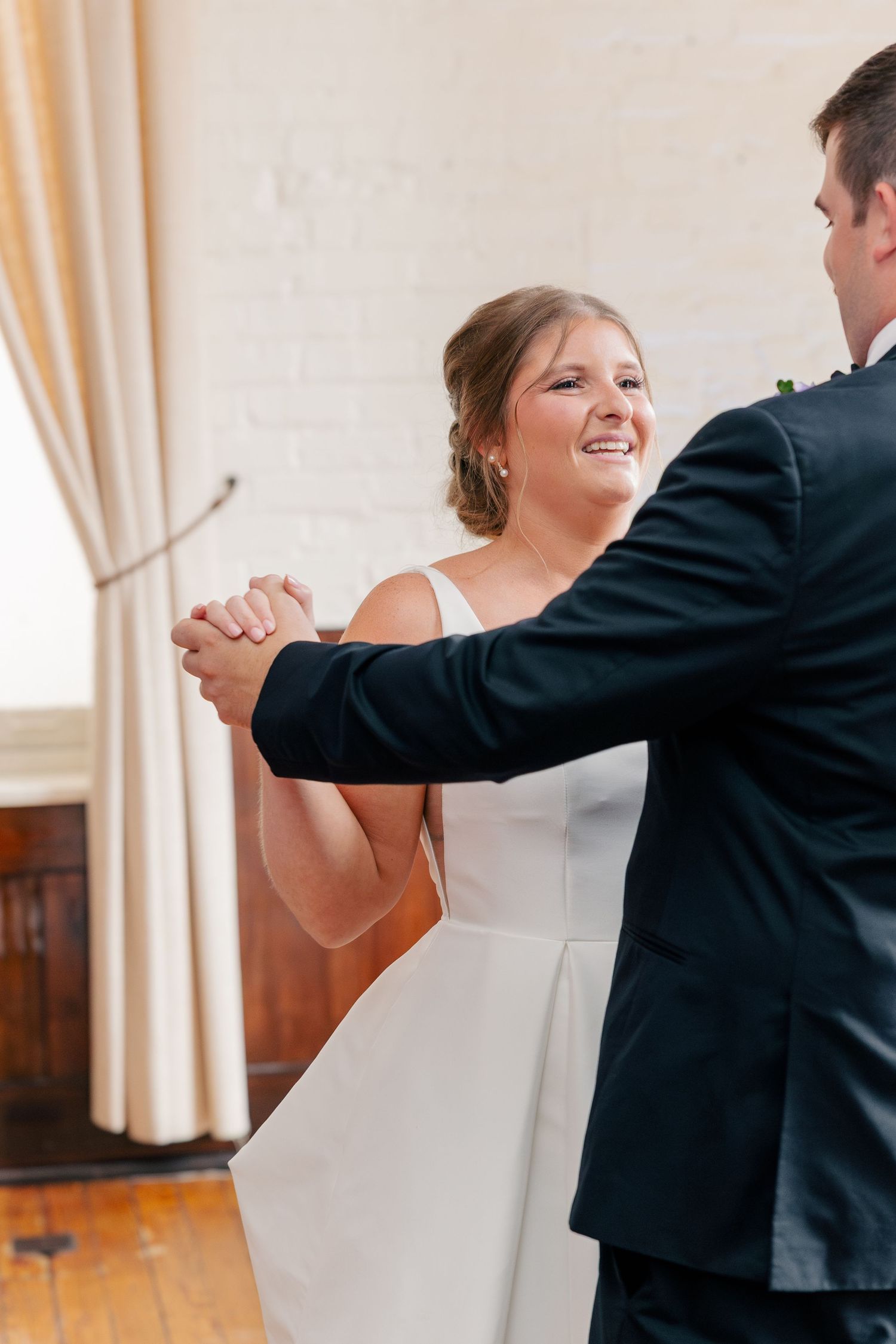 Radiant bride smiles during first dance with groom in black tuxedo in bright white venue.