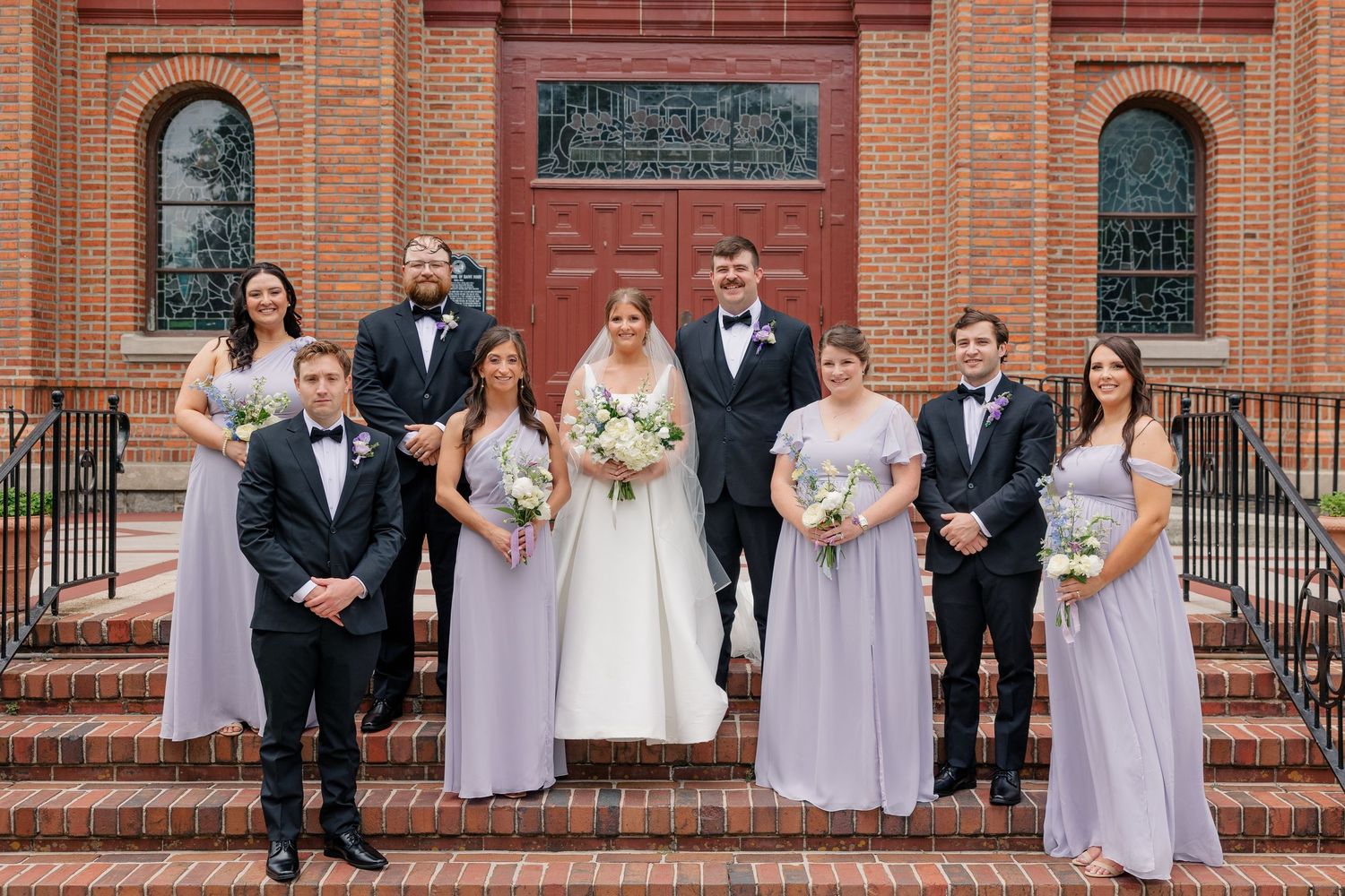 Wedding party poses on brick church steps, bridesmaids in lavender gowns and groomsmen in black tuxedos.