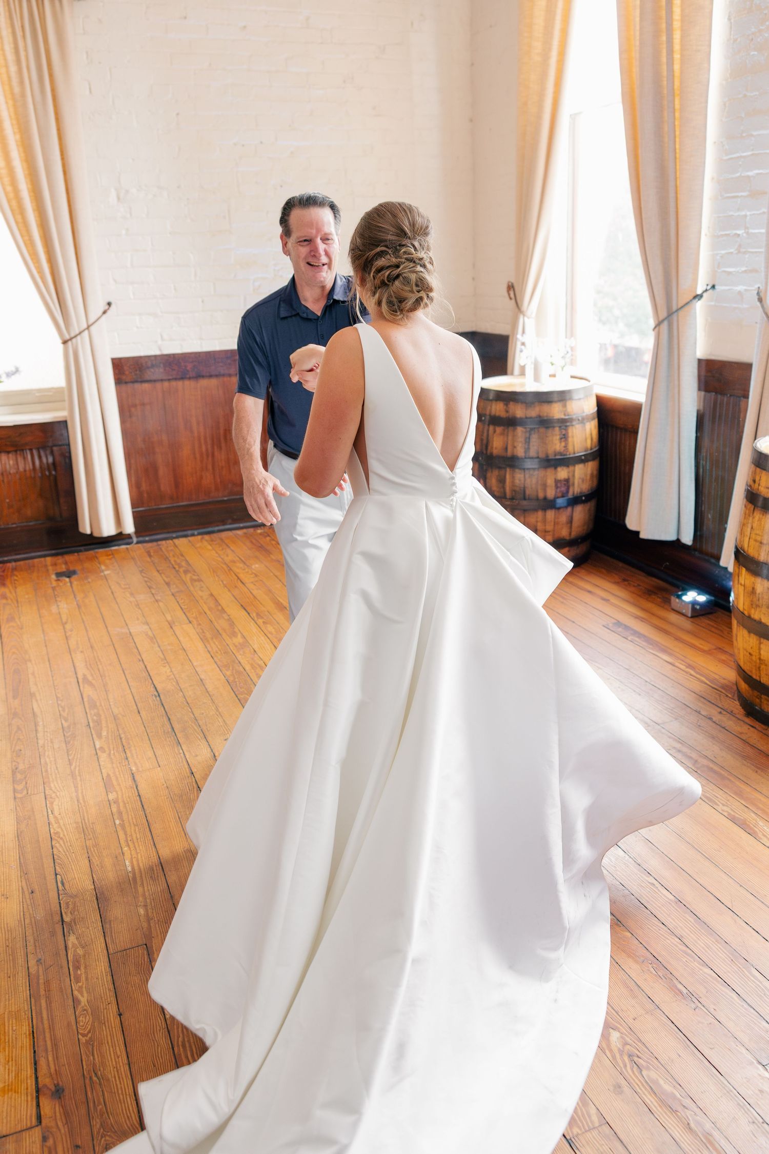 Bride in elegant white ballgown shares first look moment with smiling man in rustic wood-floored venue with barrel decor.