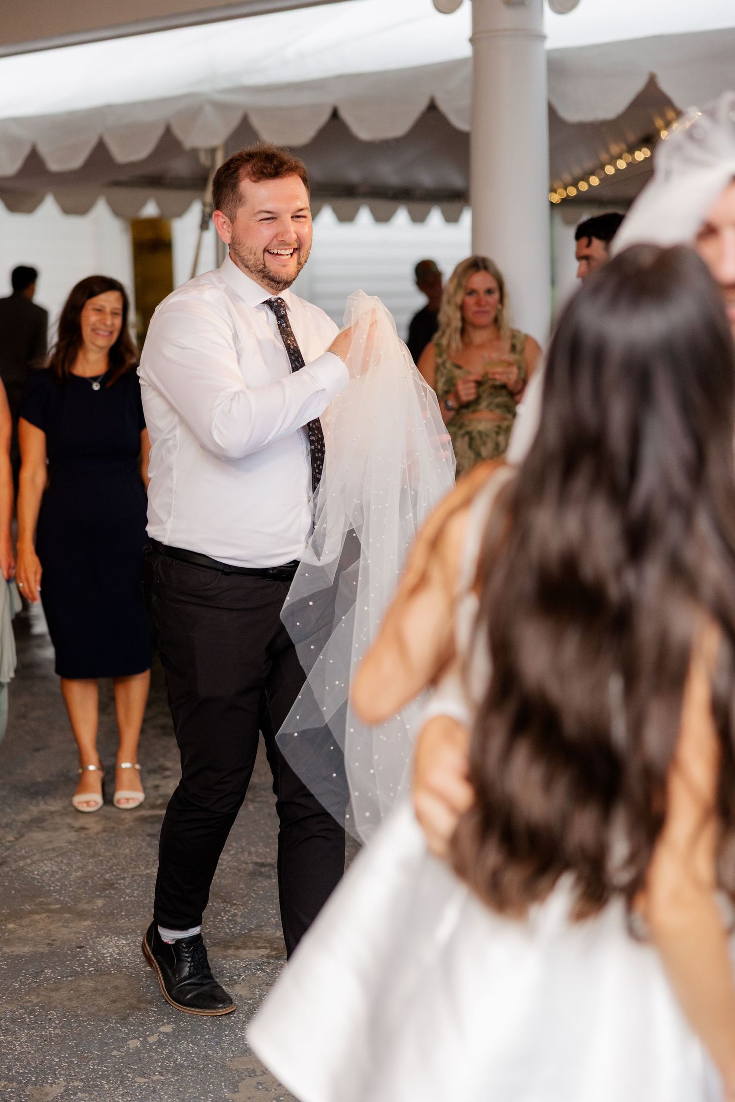 Wedding guests dance and celebrate on the dance floor during an evening reception under a white tent.