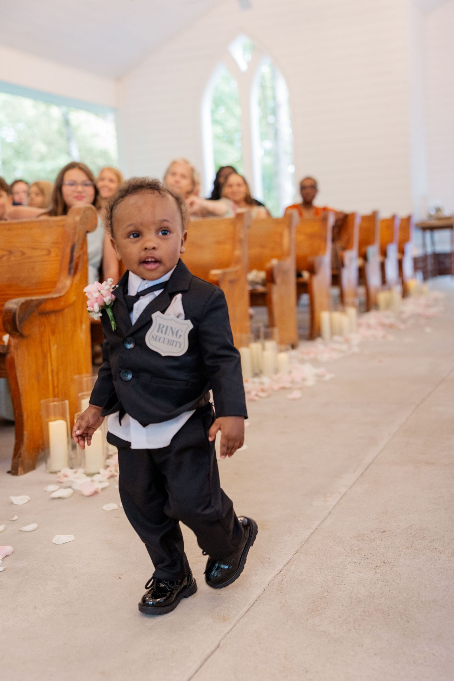 Small child in formal black tuxedo crawling down church aisle scattered with rose petals.