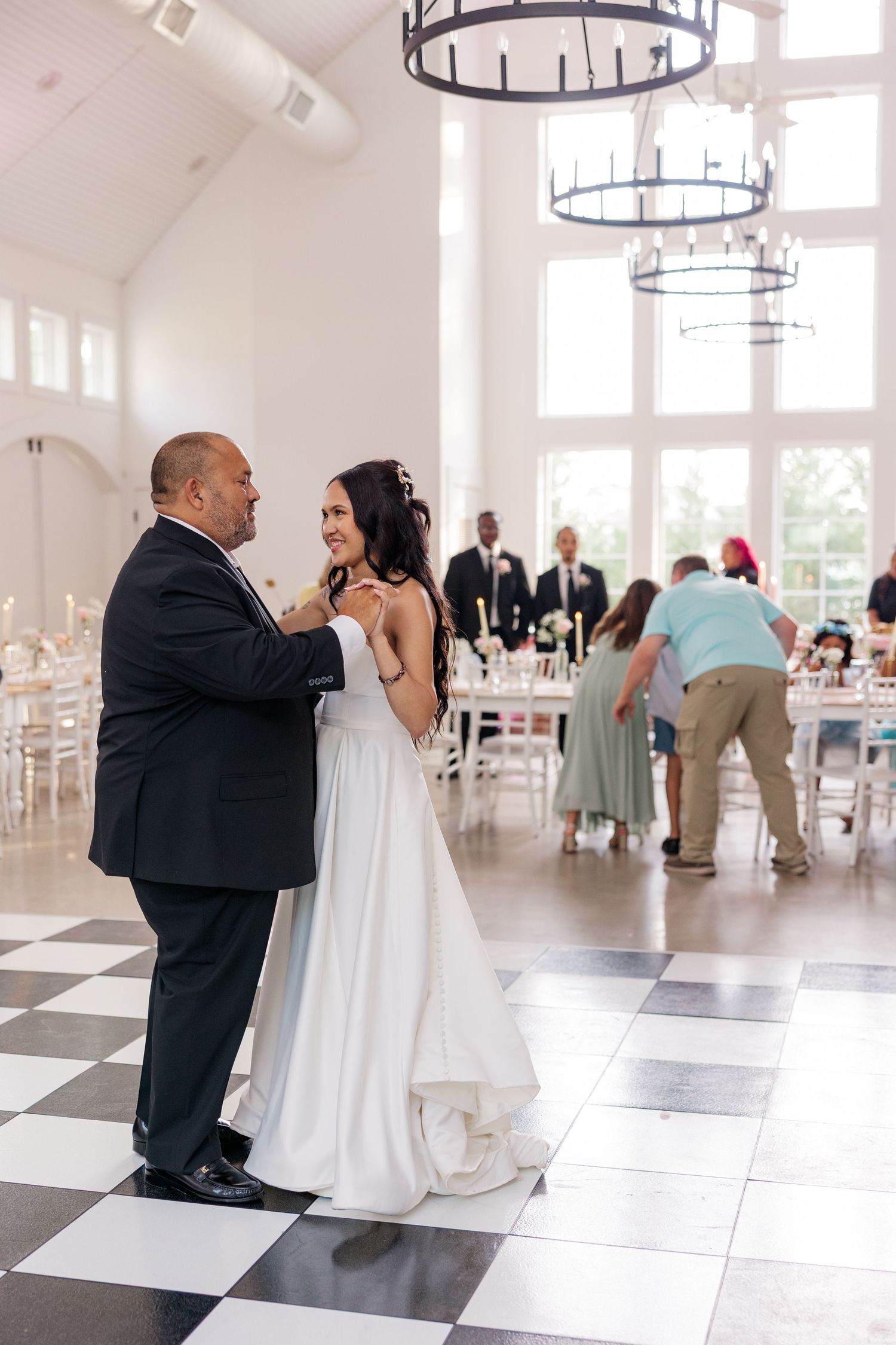 First dance moment in an elegant white reception hall with chandeliers and checkerboard floor.