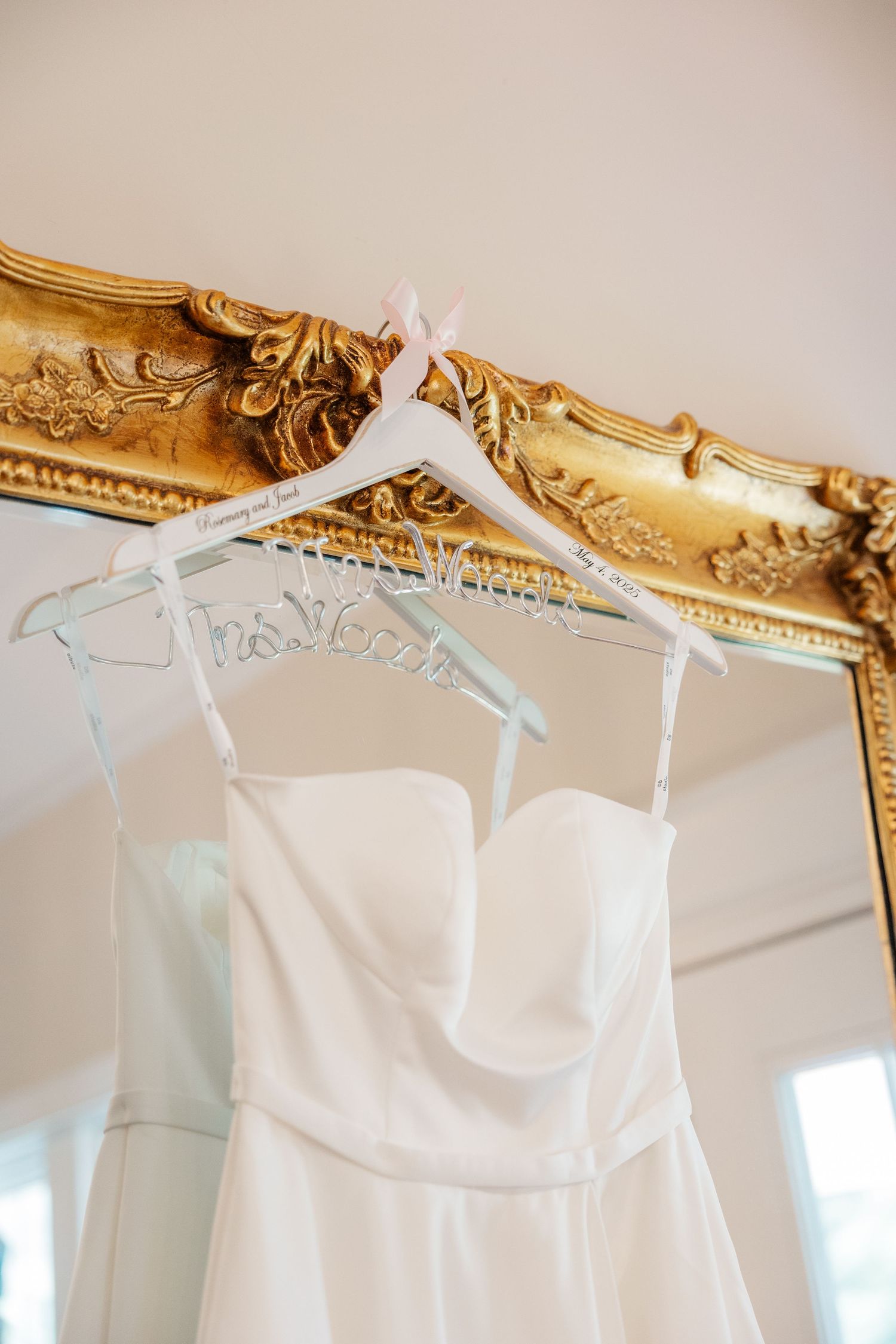 A white wedding dress on a hanger displayed against an ornate gold vintage mirror frame.
