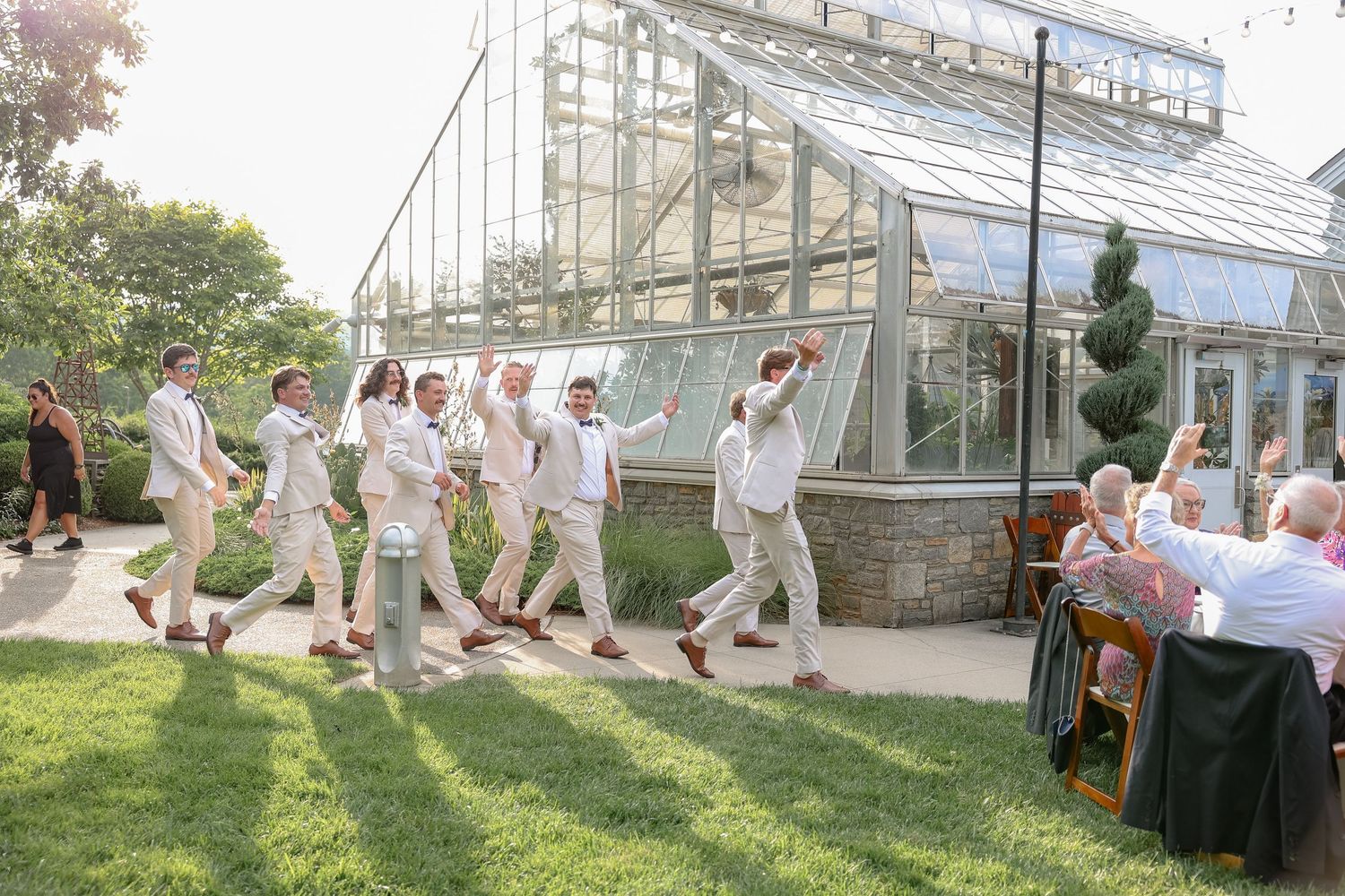 Wedding party in all-white attire dancing and celebrating outside a greenhouse venue.