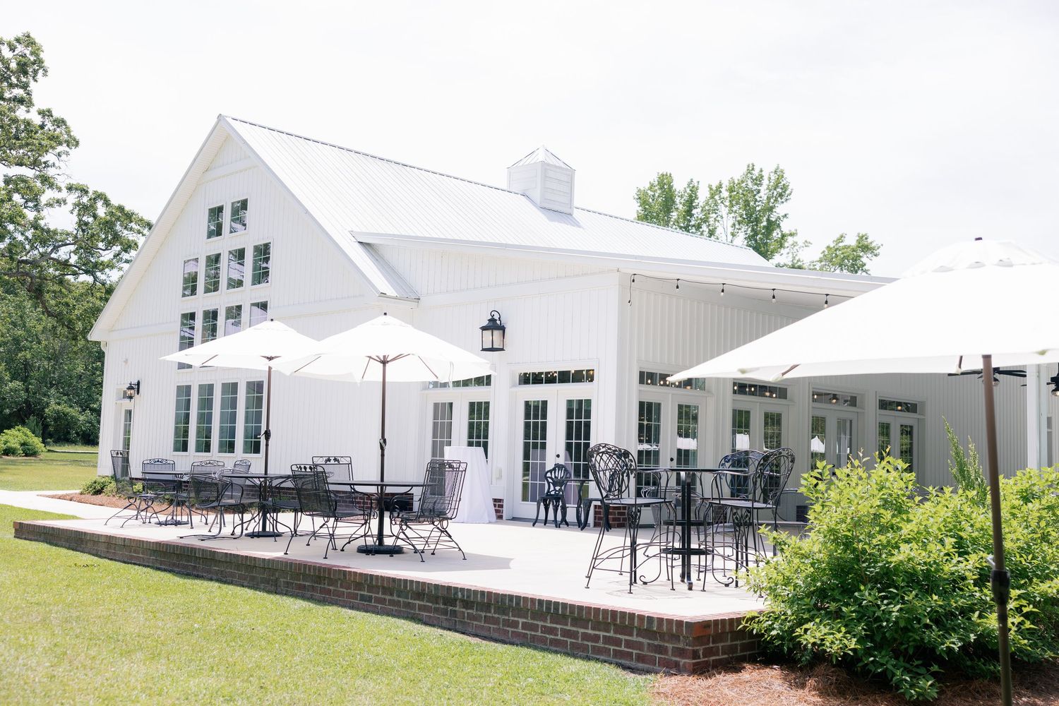 Modern white farmhouse style event venue with outdoor patio seating and landscaping against green lawn background.