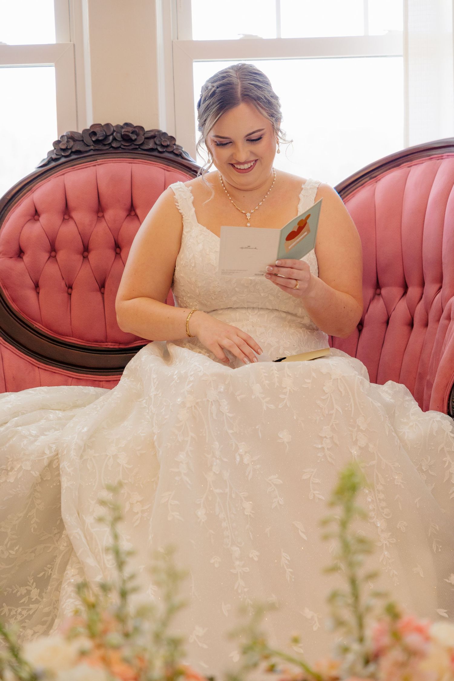 A bride in a white lace dress sits on a pink vintage couch reading something.