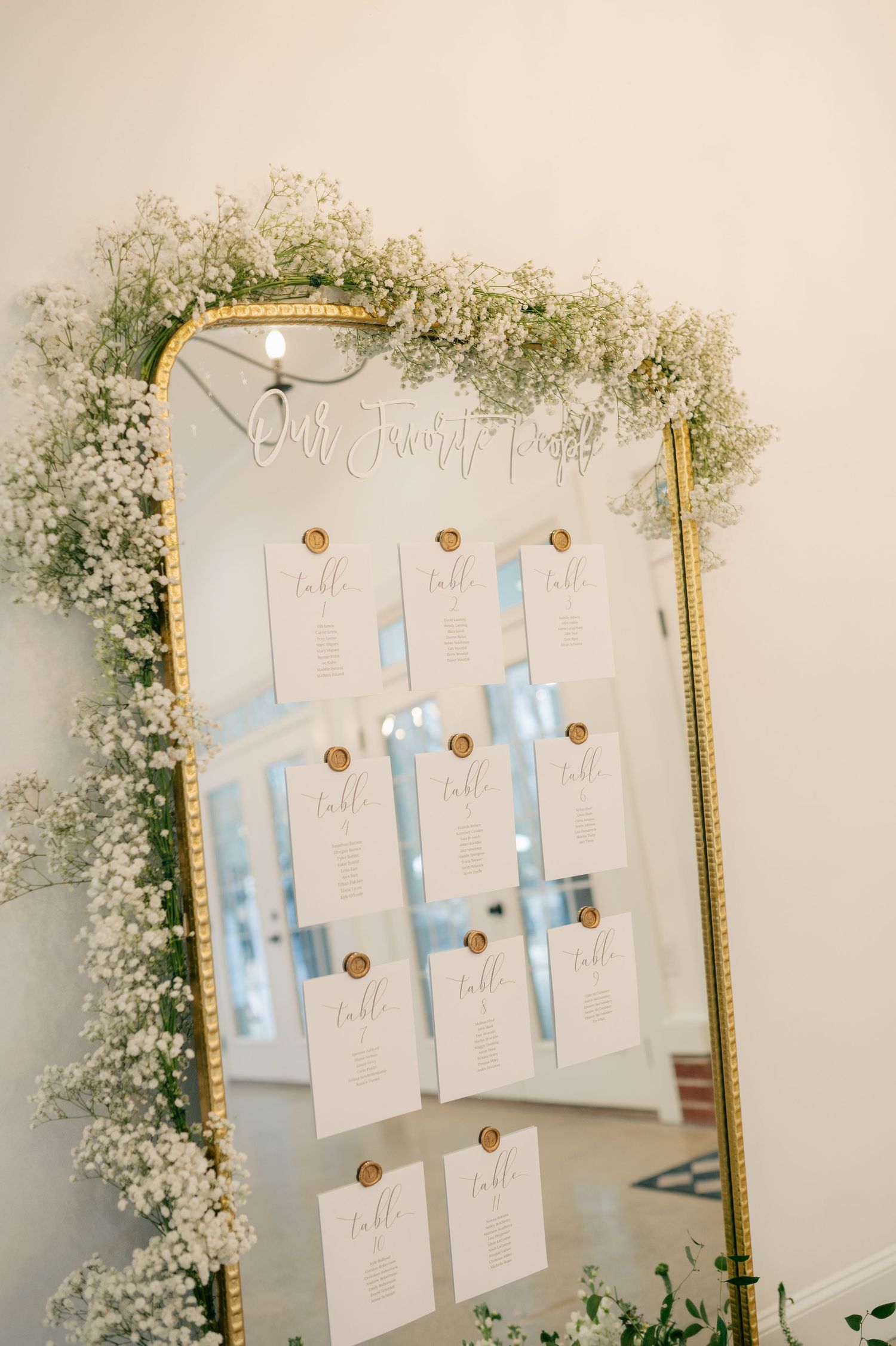 A gold-framed mirror decorated with baby's breath displays wedding seating arrangements.