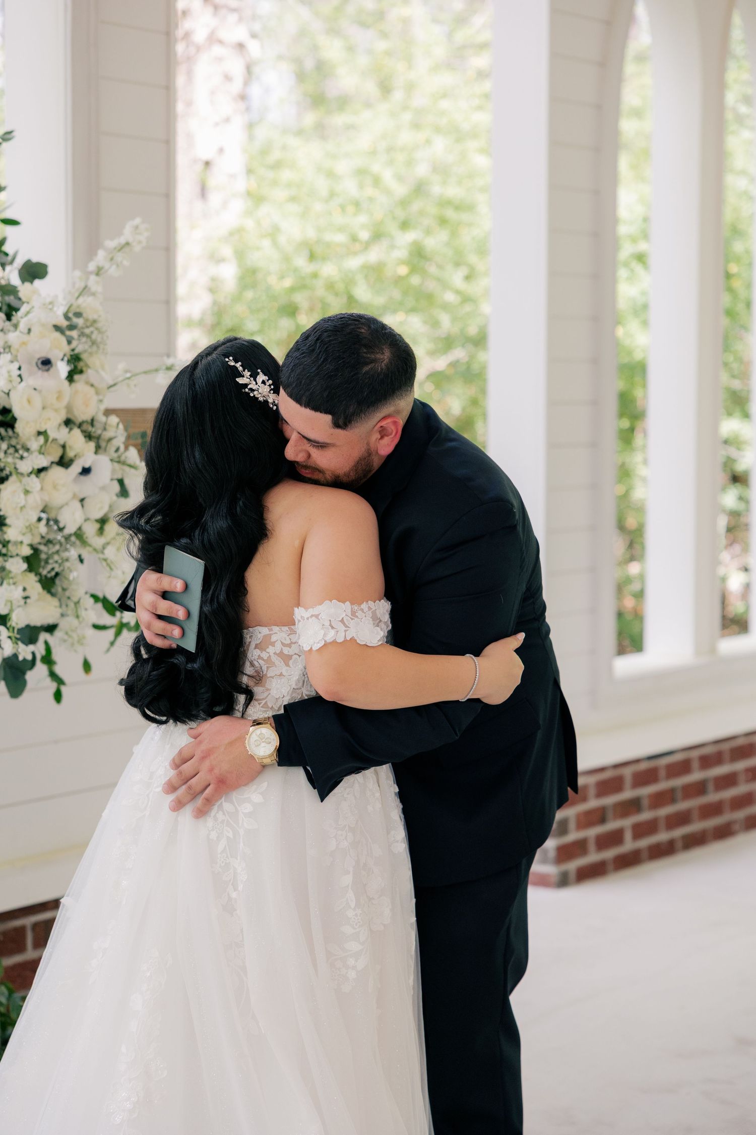 Couple embraces during first look moment on white columned porch with floral backdrop.