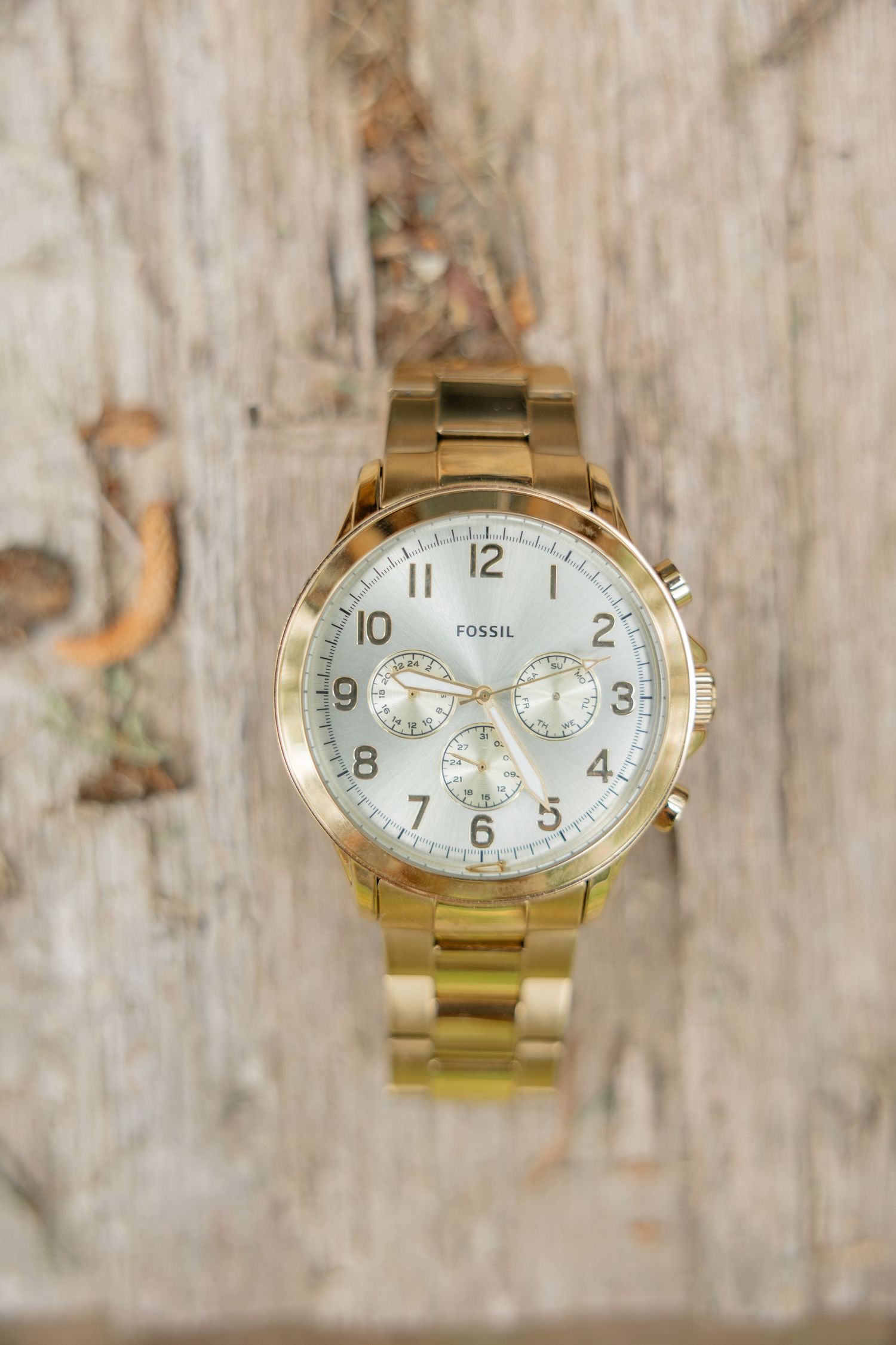 Gold men's Fossil watch with chronograph features displayed at different angles on a textured wooden surface.