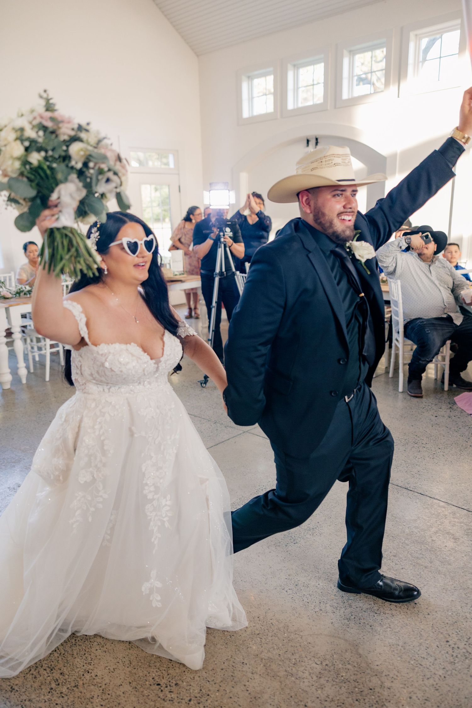 Wedding party dancing and celebrating in a bright indoor reception venue with white decor.
