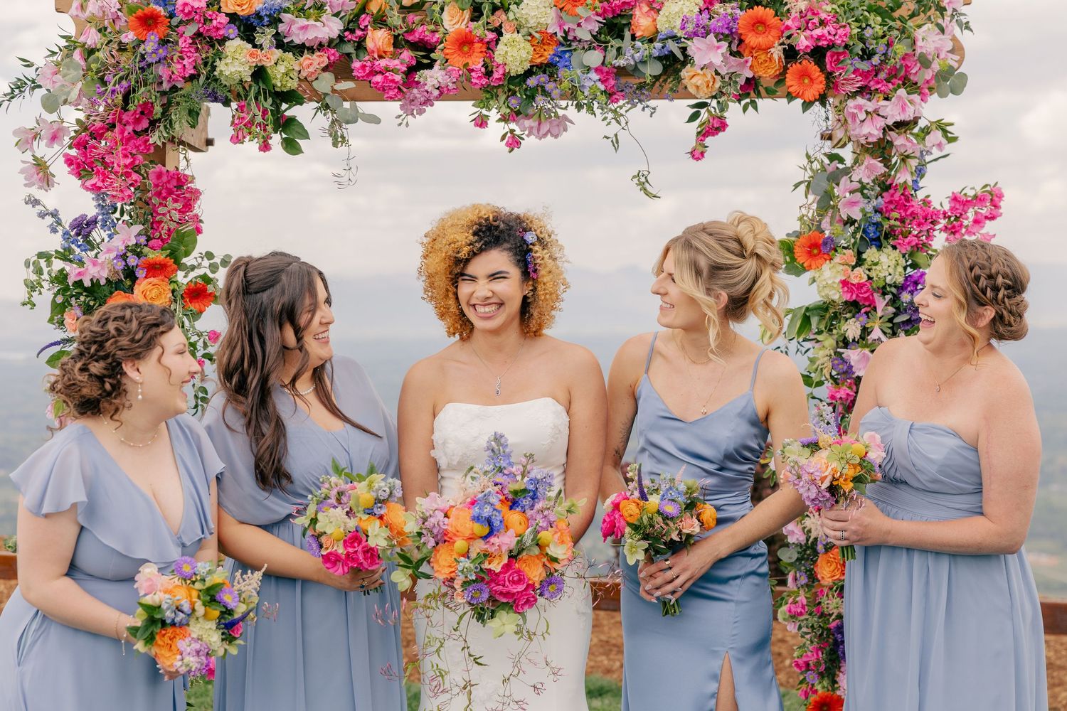 Bride with bridesmaids in blue dresses holding colorful bouquets under floral arch.