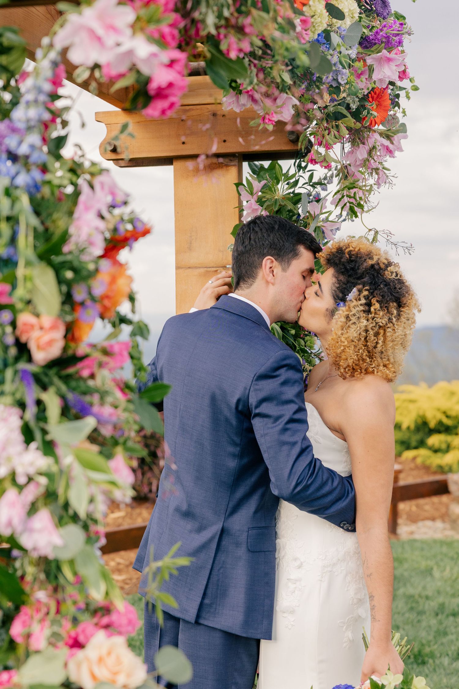 Bride and groom share first kiss under floral arch adorned with colorful flowers at outdoor wedding.