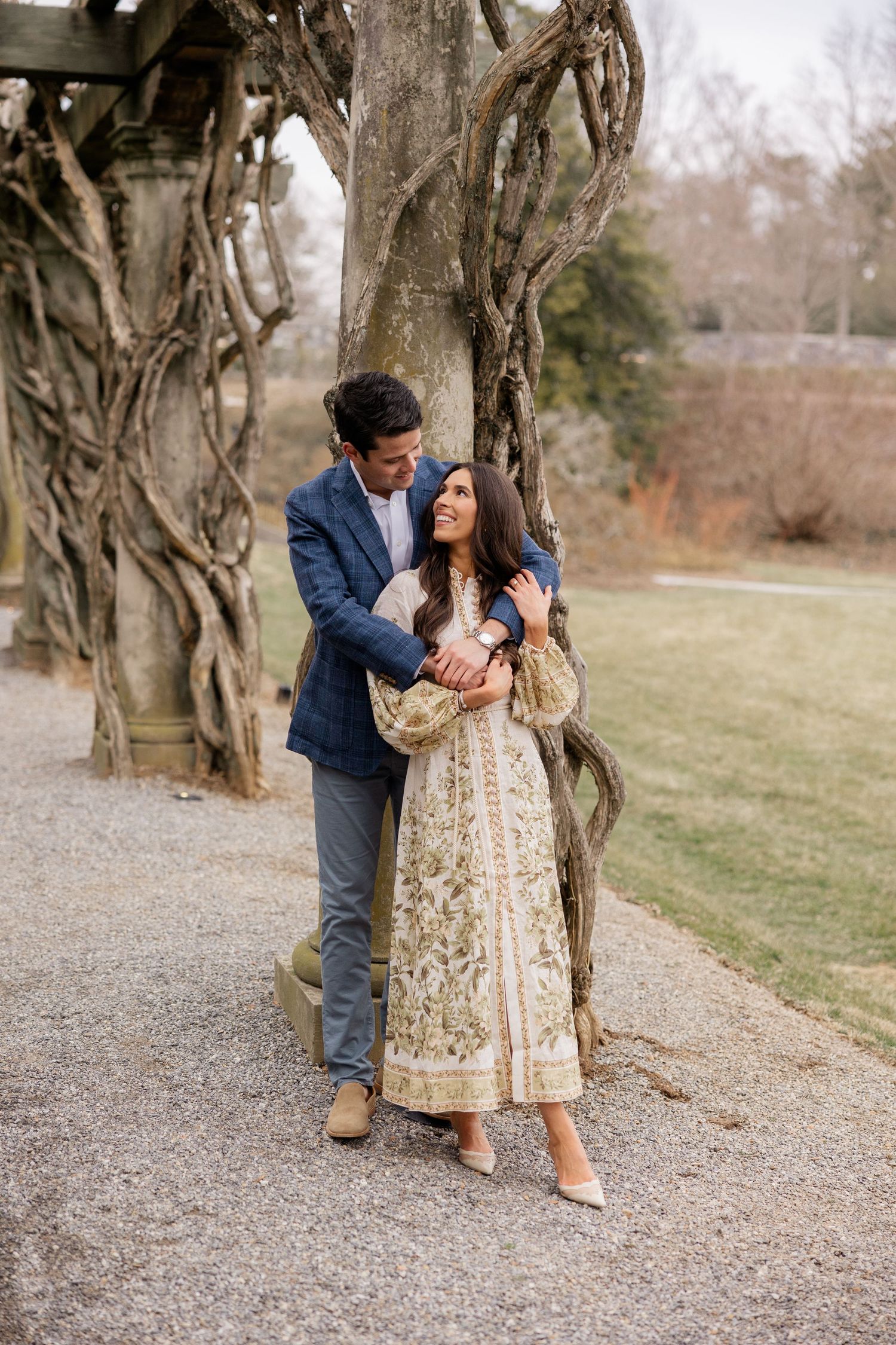 A couple embraces on a gravel path under twisted tree branches in a romantic outdoor setting.