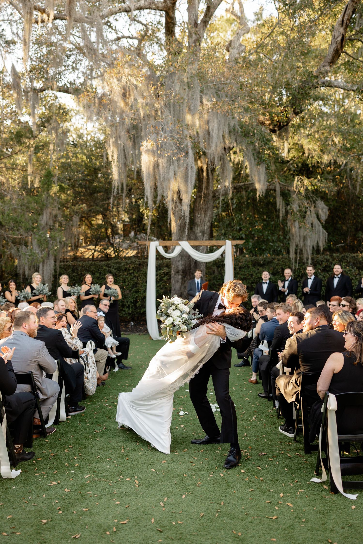 A romantic dip during a wedding ceremony under mossy oak trees as guests look on from rows of black chairs.