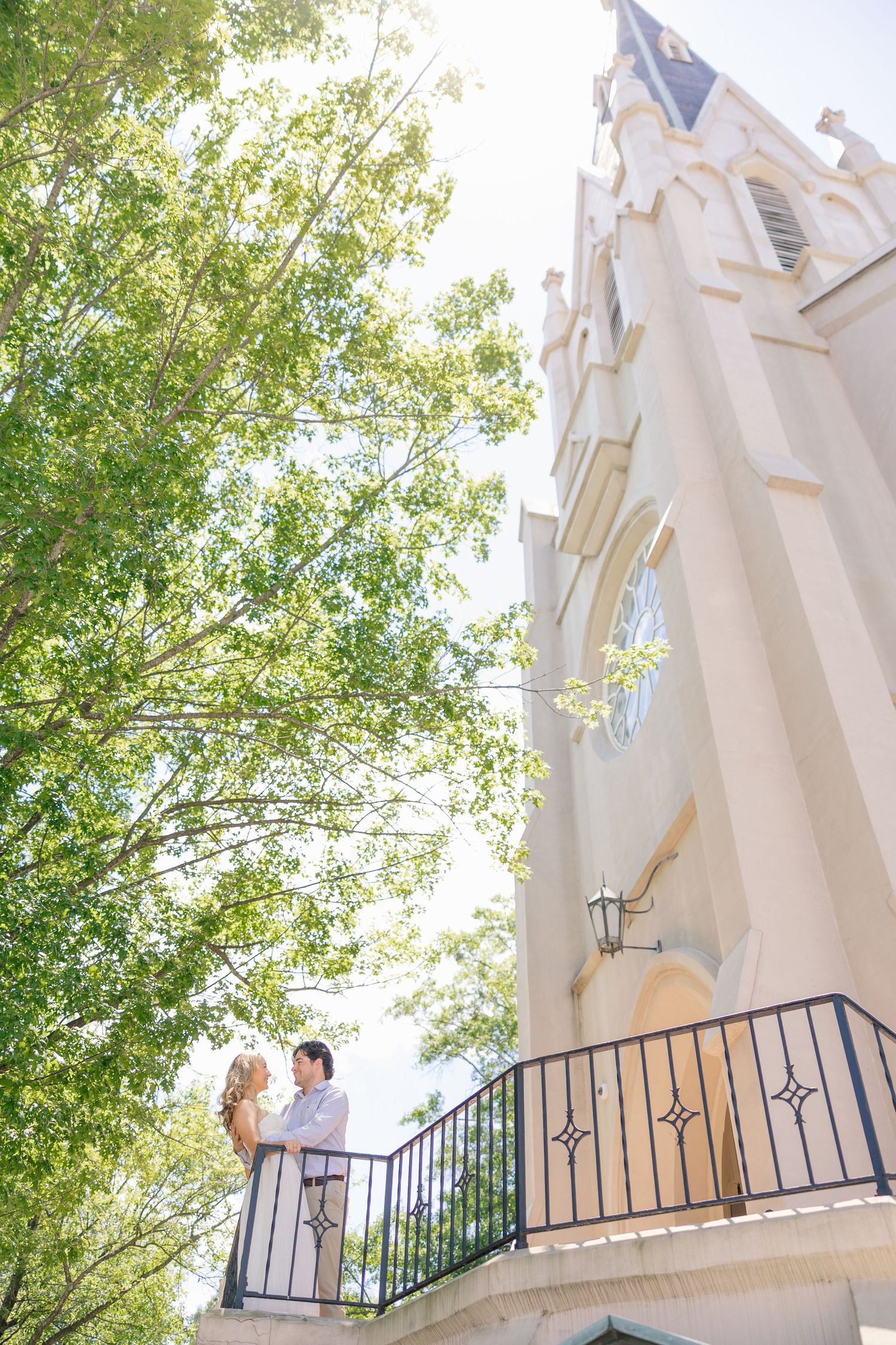 Couple embracing on a balcony beside a white church tower with green trees on a bright day.