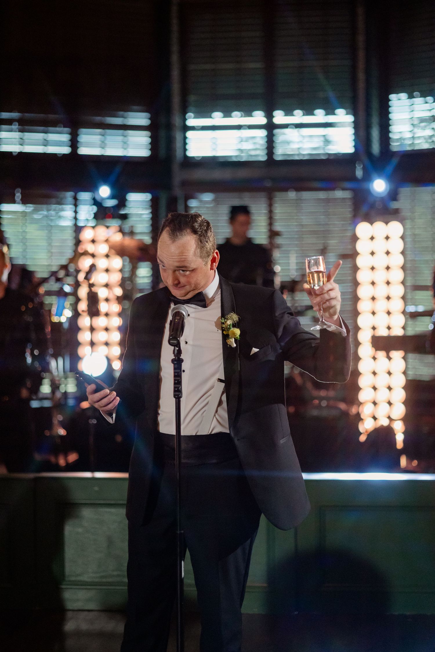 Man in tuxedo raises champagne glass while giving a wedding toast at a stylish reception venue.
