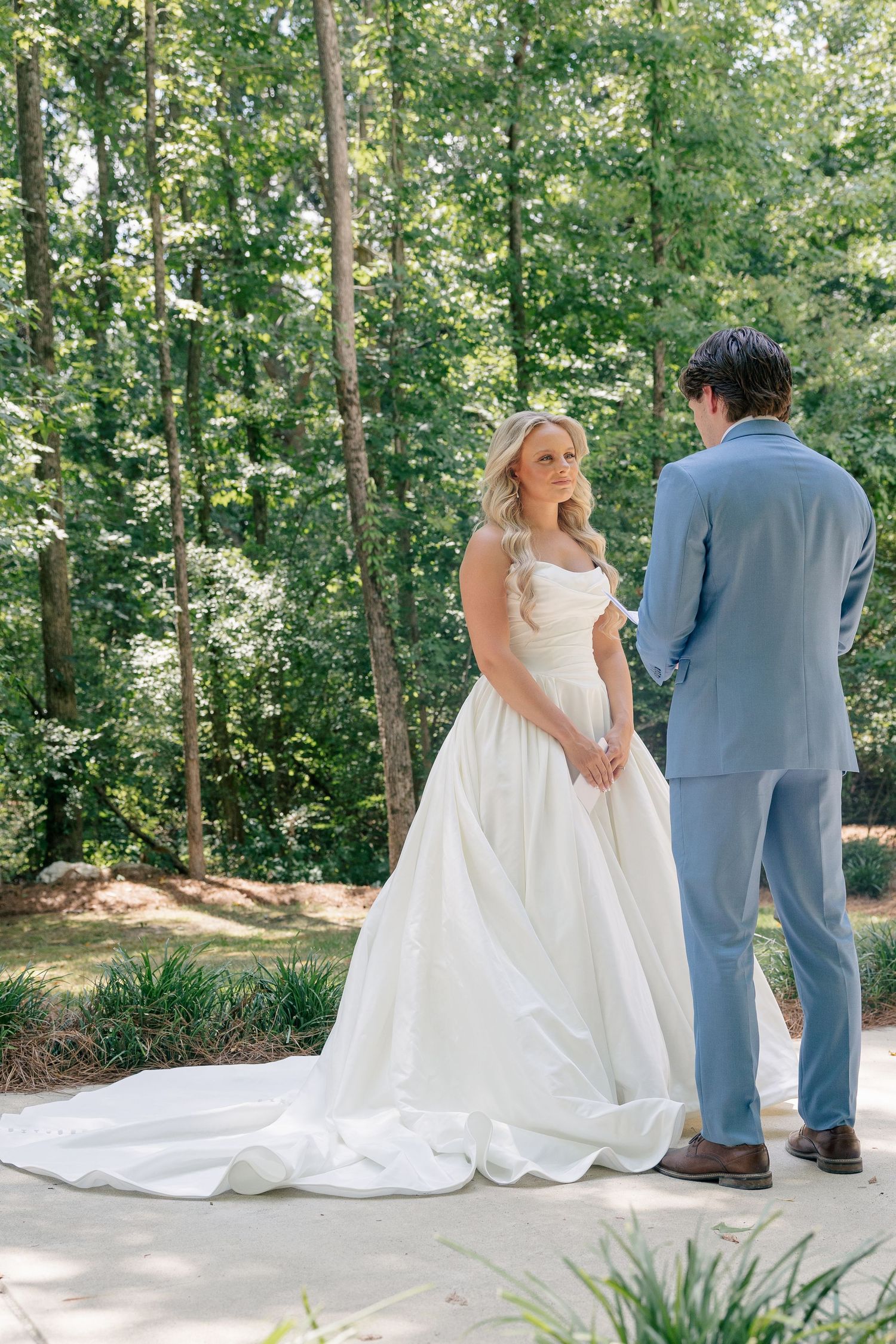Bride in white ball gown and groom in light blue suit share a first look outdoors in a lush green forest setting.