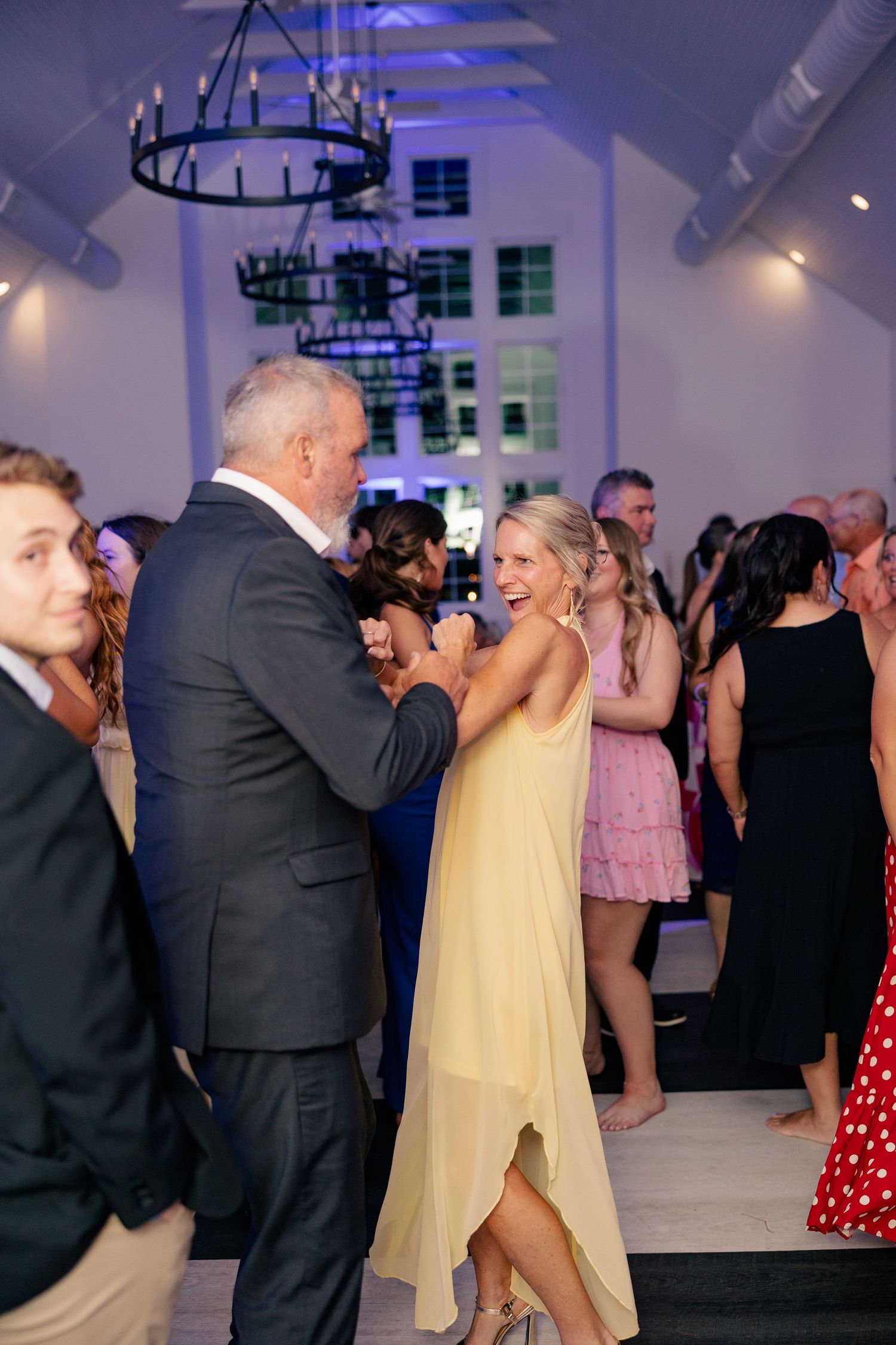 Guests dancing and laughing at a wedding reception under a chandelier in an elegant white venue.